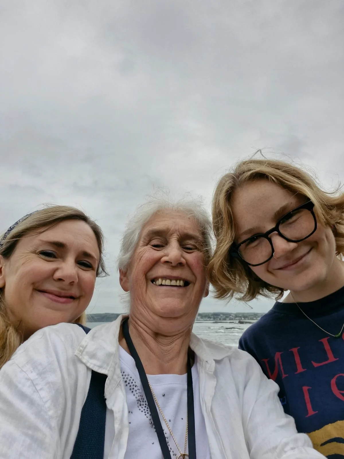 Three smiling women taking a selfie outdoors on a cloudy day, with water and a distant shoreline in the background.