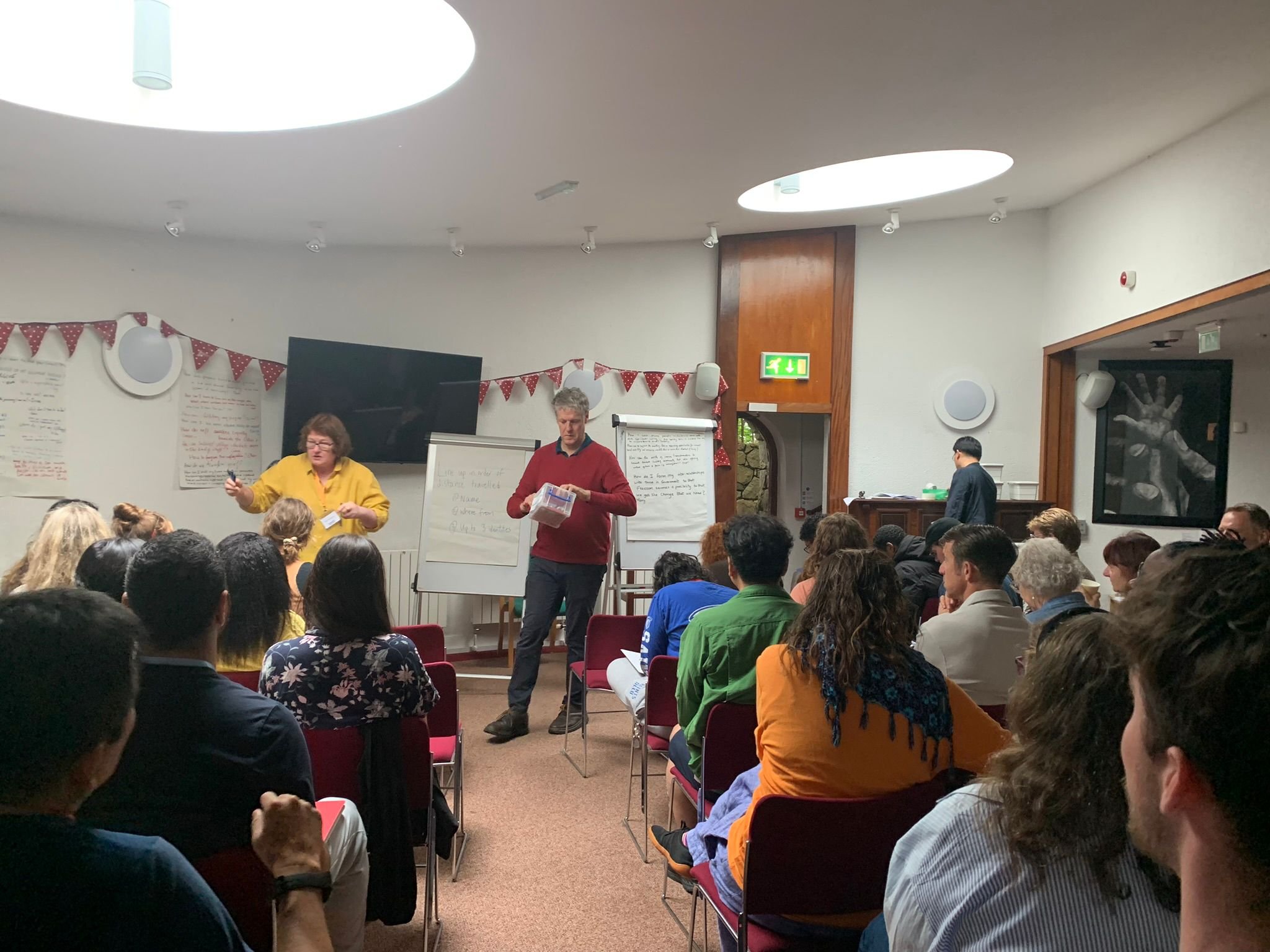 A classroom or conference room filled with people attending a presentation or workshop. Two presenters are at the front, one holding a marker and the other holding a clear plastic container. There are flip charts, a television, and red triangular ban