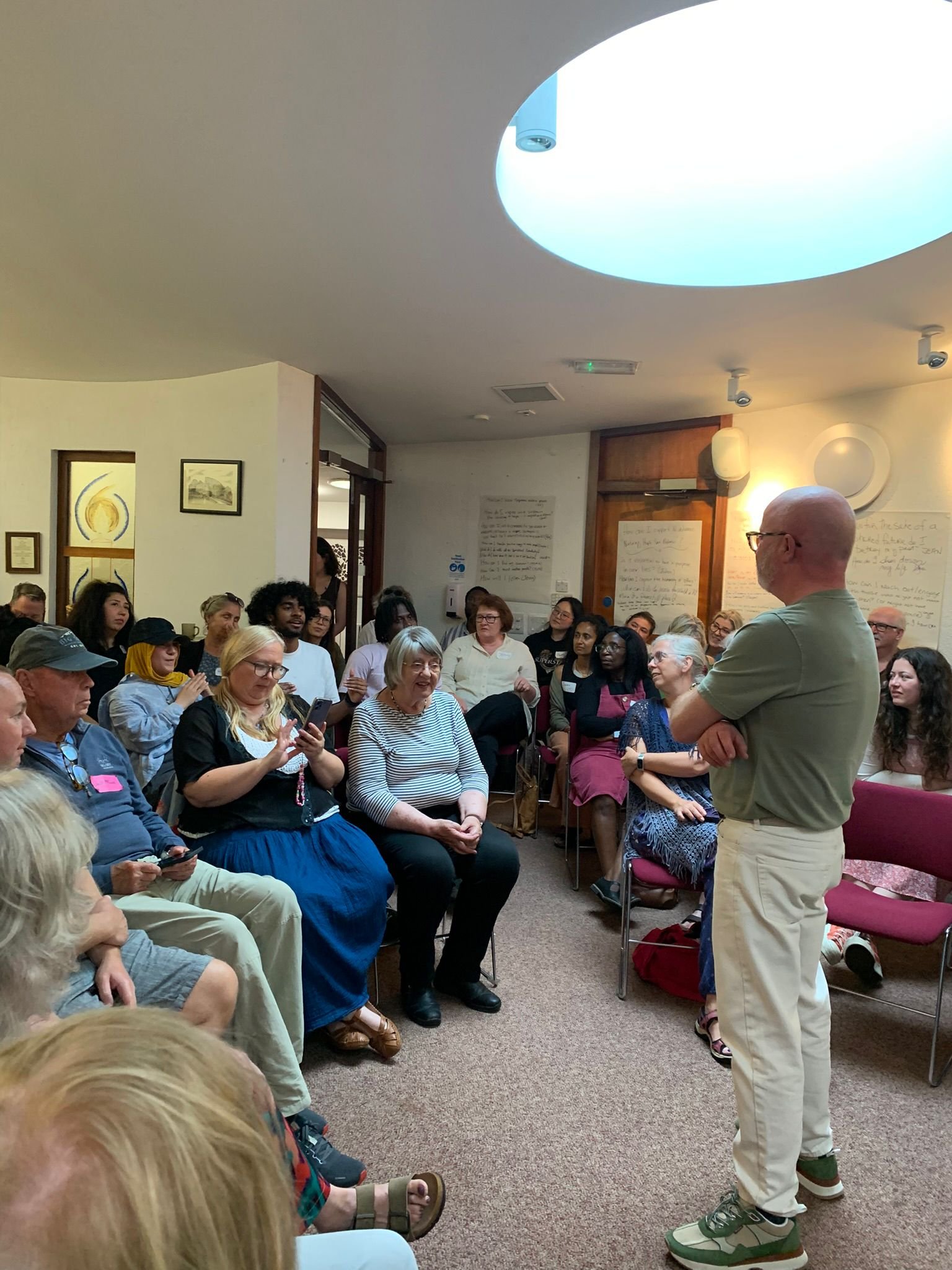 A group of people sitting and listening to a man speaking in a room with white walls and artwork, some people are taking notes or photos, and a woman in front is smiling and looking at her phone.