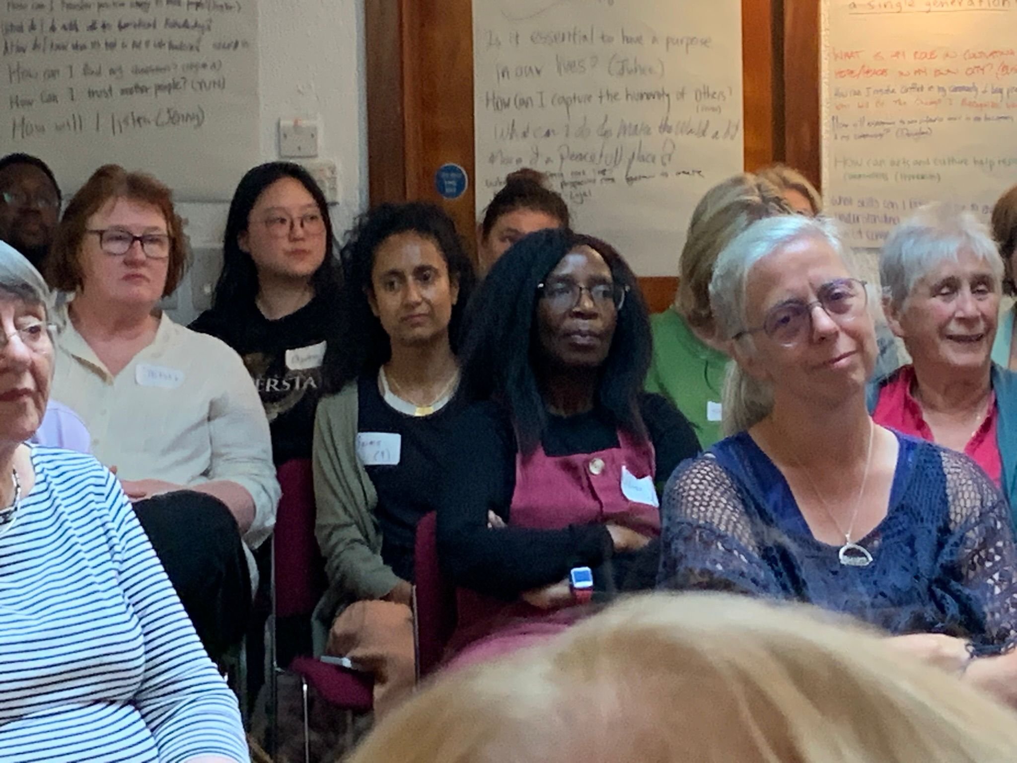 A diverse group of women seated and attentively listening at an indoor event, with handwritten notes on posters on the wall behind them.