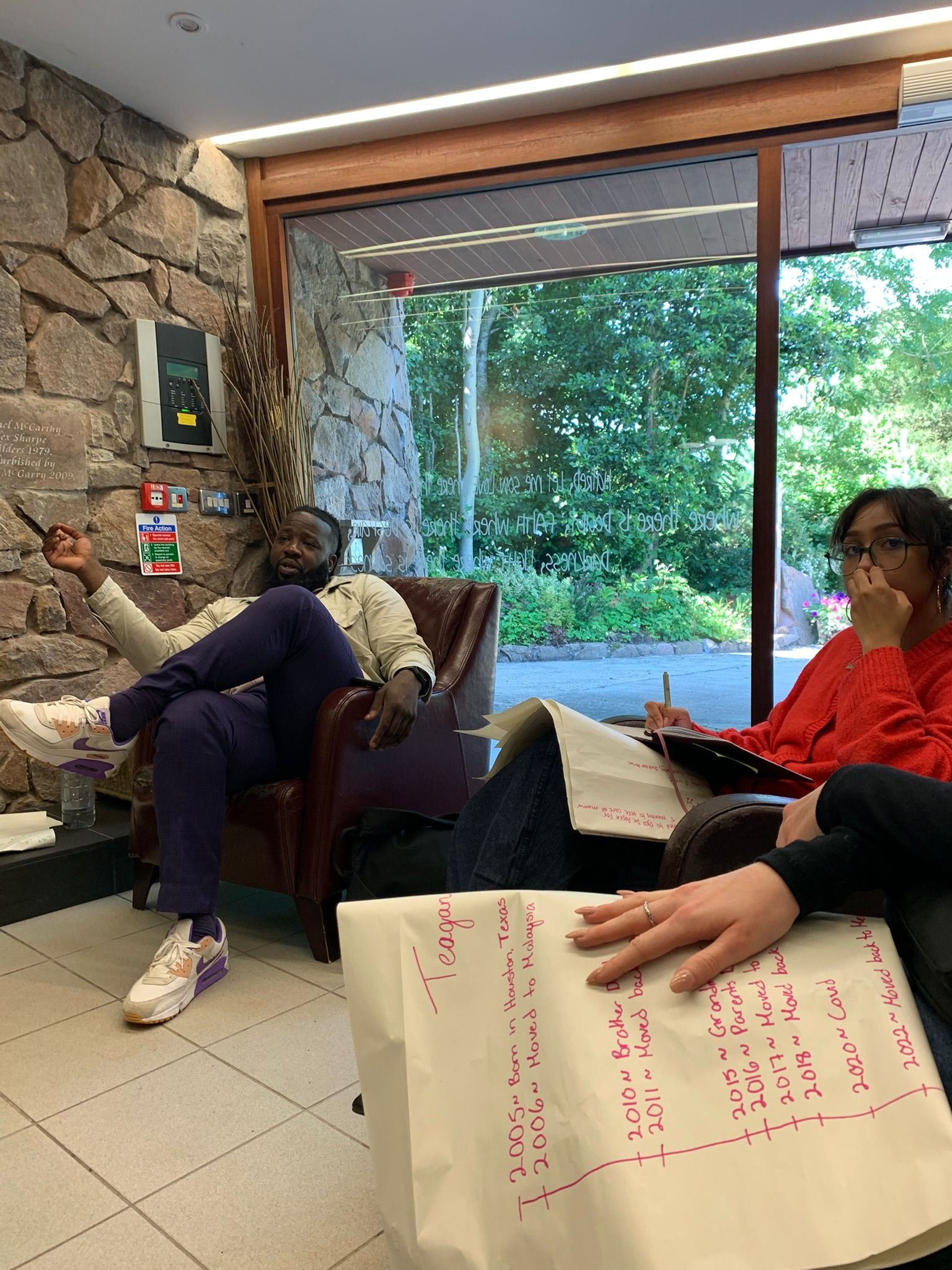 Two people sitting inside a coffee shop or café, near a large window showing outdoor greenery. One person is sitting in a leather armchair, gesturing with their hand, while the other is taking notes with a pen in a notebook, with handwritten notes on