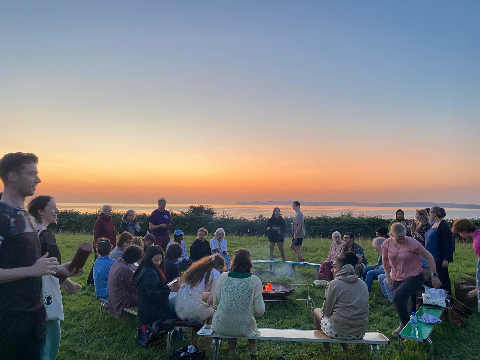 A group of people gathered outdoors near a body of water during sunset, with the sky displaying warm colors and some standing while others sit around a small campfire.