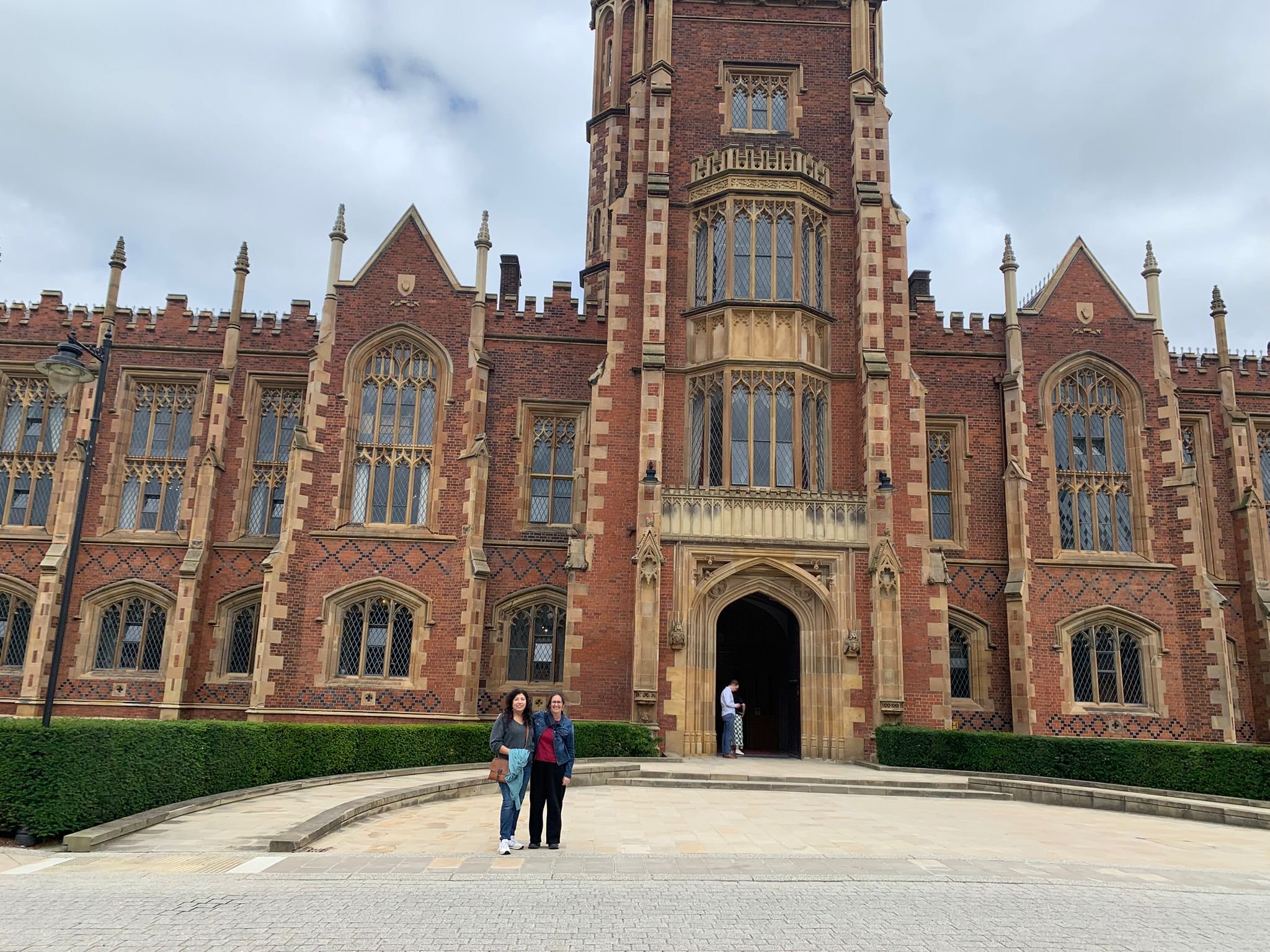 Two women standing in front of a historic brick building with ornate windows and a large entrance, with a person near the door and a cloudy sky above.
