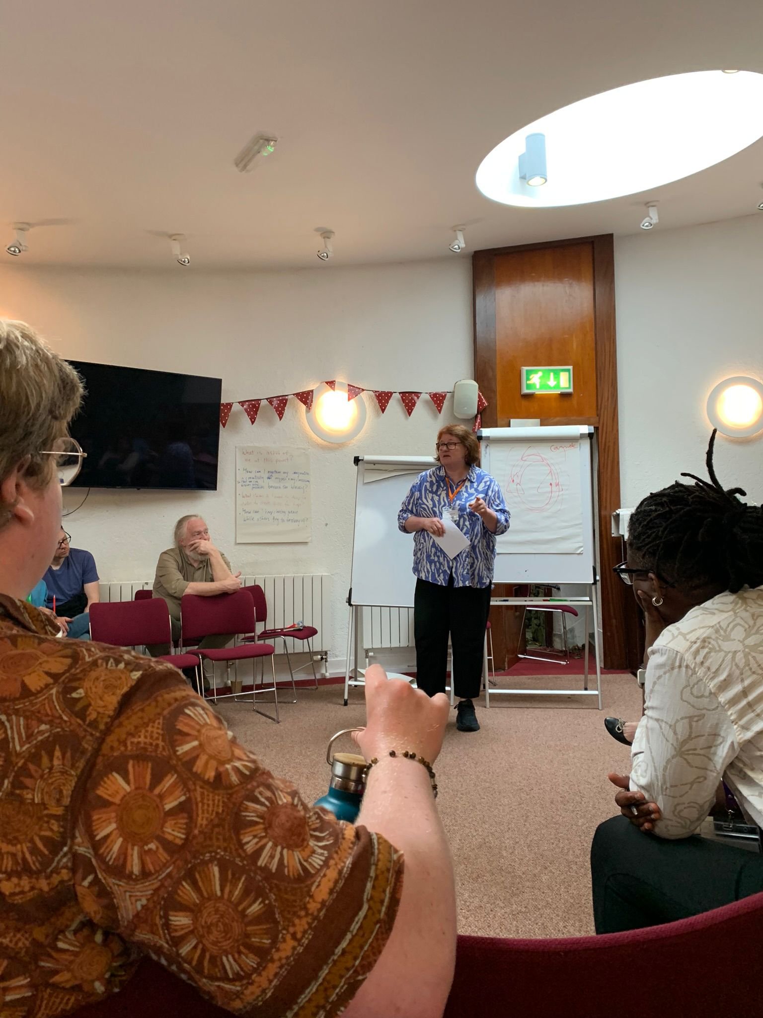 A woman standing and speaking to a group in a conference room with a whiteboard behind her, a television on the wall, and a decorative garland.