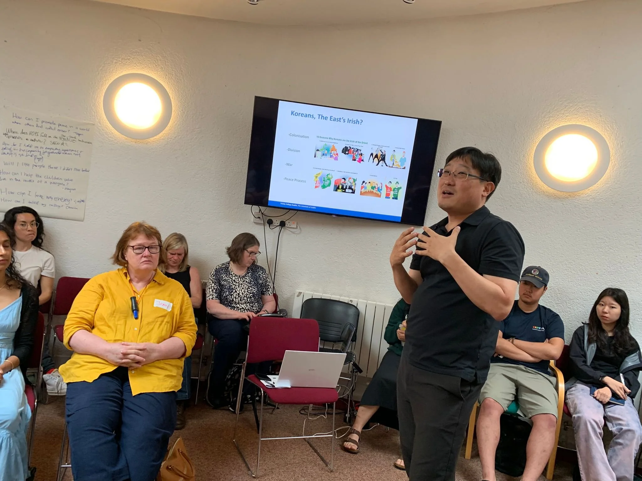 A man giving a presentation to an audience in a conference room. The presentation slide on the screen behind him discusses Koreans and the East's Irish, with illustrations and bullet points. The audience members are seated and listening attentively.