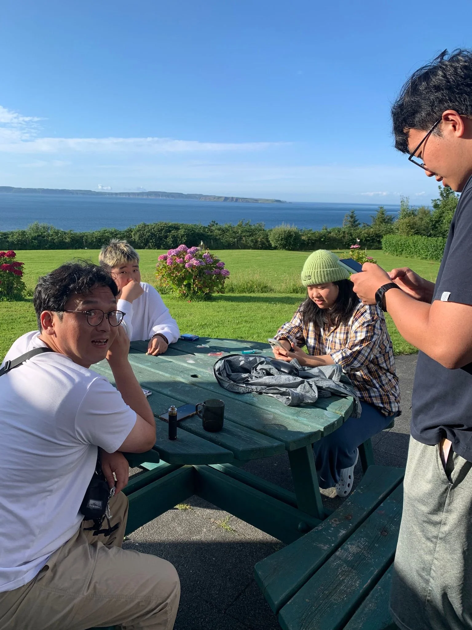 Four people sitting and standing around a green picnic table outdoors, with a view of the ocean and blue sky in the background. One person is looking at the camera with a surprised expression, while the others are focused on their phones.