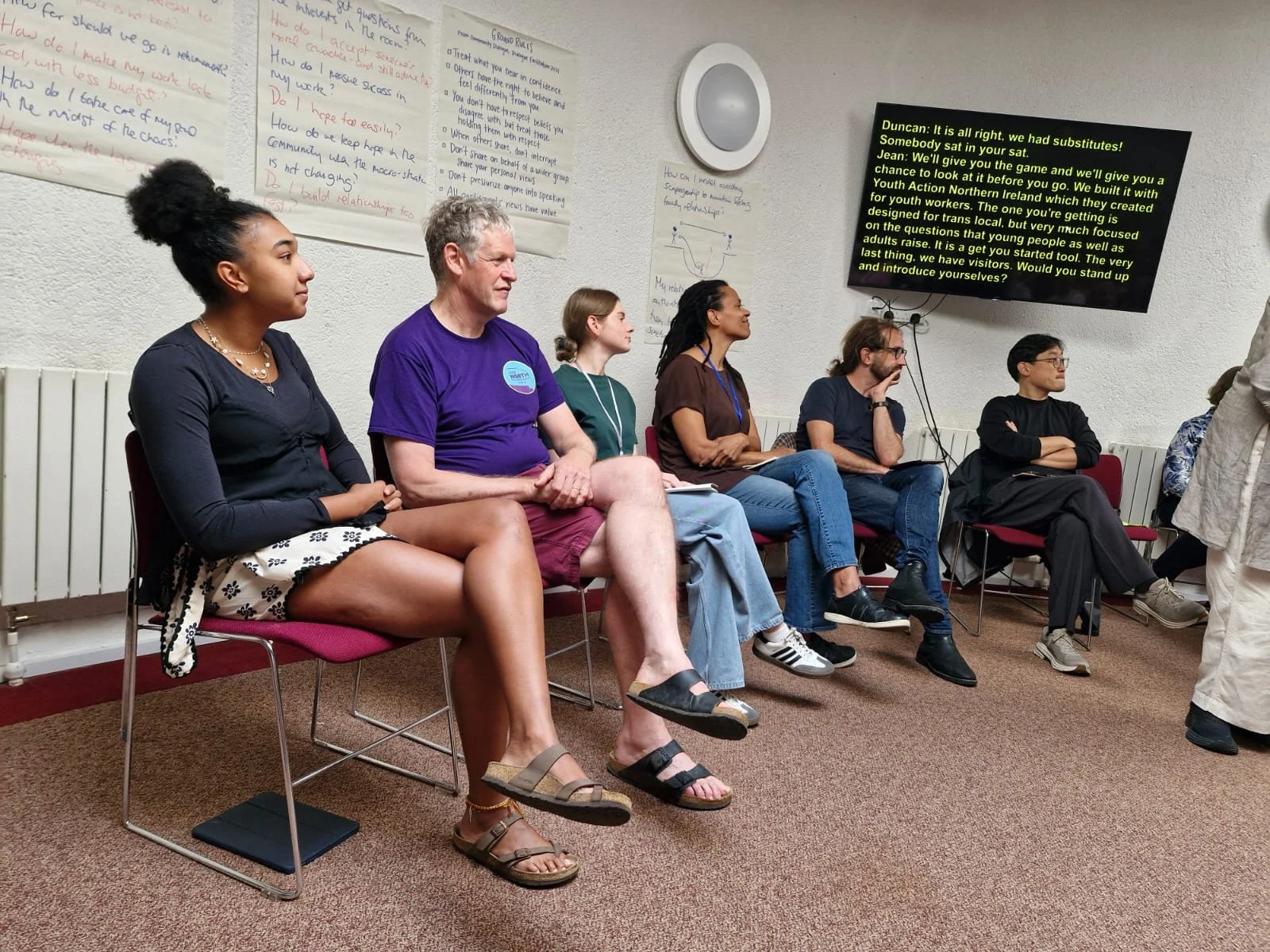 A group of six people sitting in a row of chairs in a classroom or workshop setting, with whiteboards and a large screen displaying text in the background.