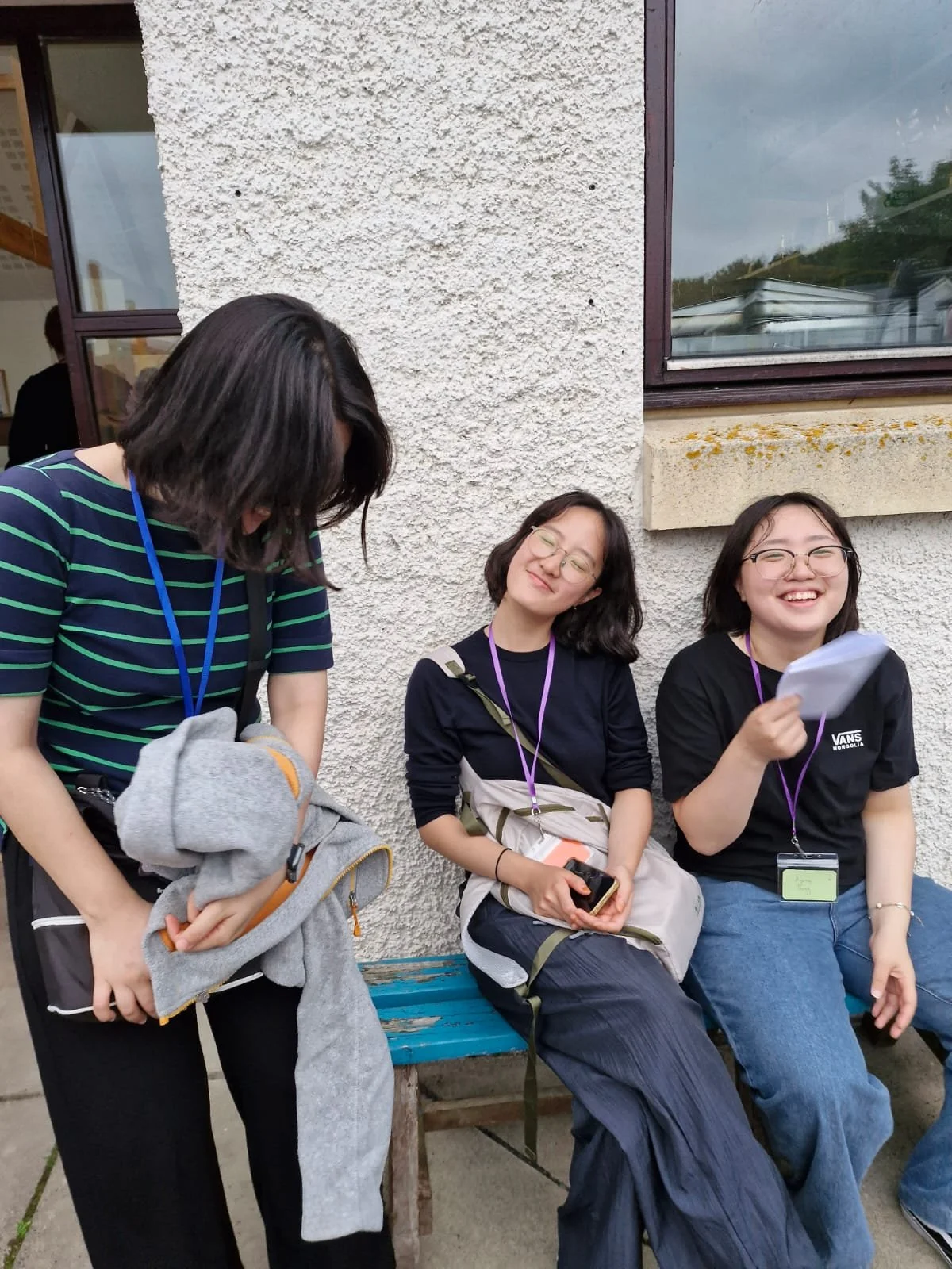 Three young women sitting outside against a textured beige wall, laughing and smiling. One woman on the left is standing, looking down and holding a gray jacket. The woman in the middle is sitting with her eyes closed, smiling contentedly, holding a 