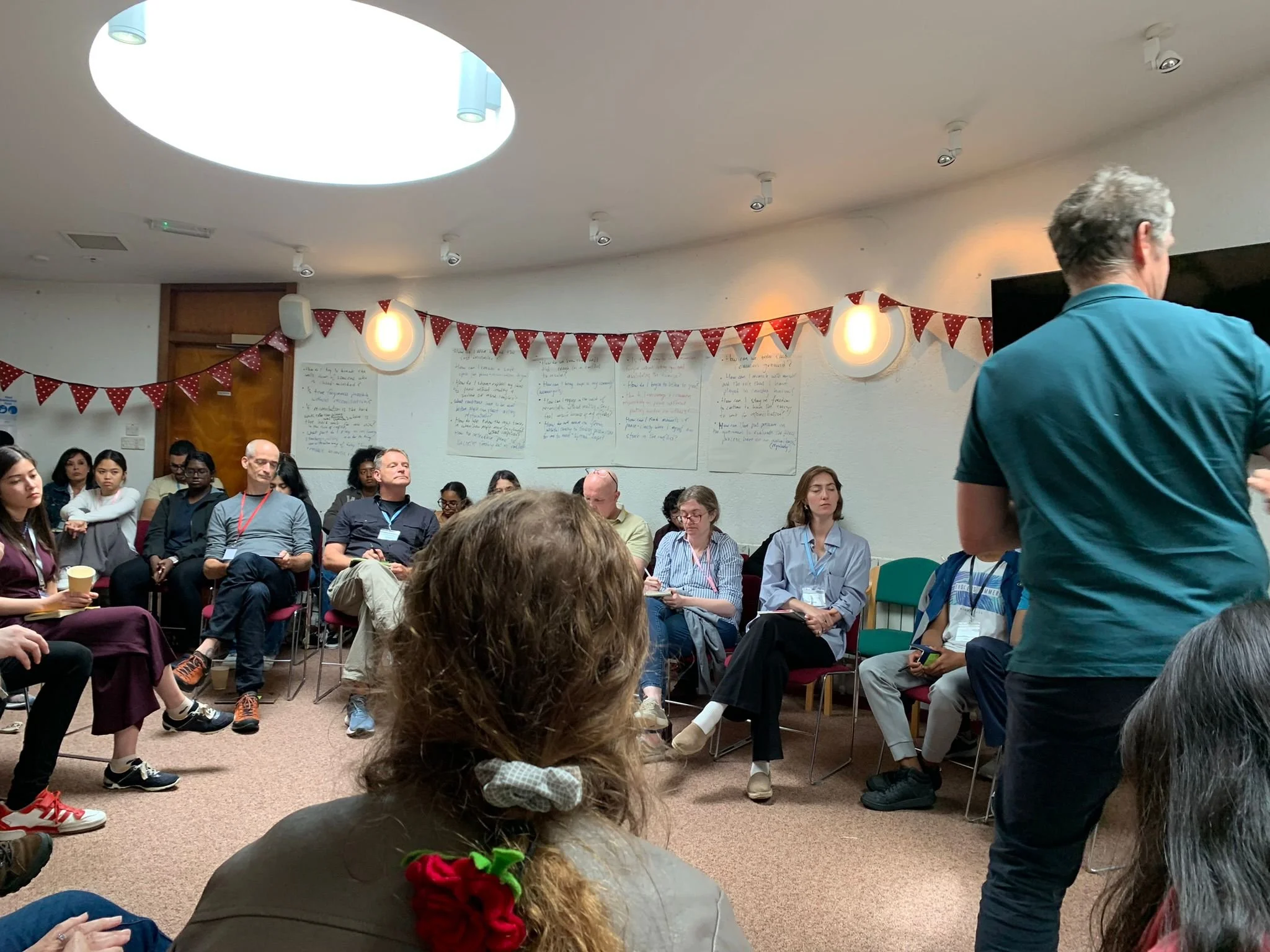 A group of people sitting in a circle in a conference room, listening to a speaker who is standing on the right side with his back to the camera. The room has white walls, posters with handwritten notes, and red bunting decorations.