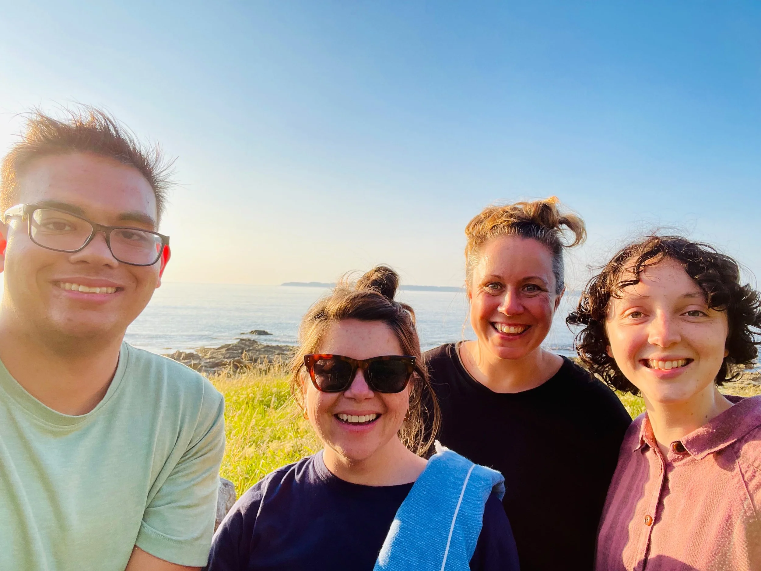 A group of five smiling people taking a selfie outdoors near a body of water, with a clear blue sky in the background.