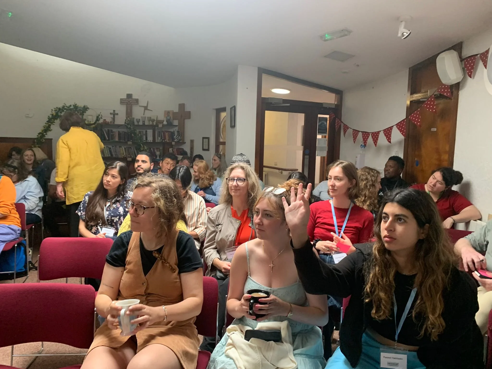 A group of diverse people sitting in a room, some raising hands, others holding cups, attending a meeting or conference with one woman in the foreground raising her hand.