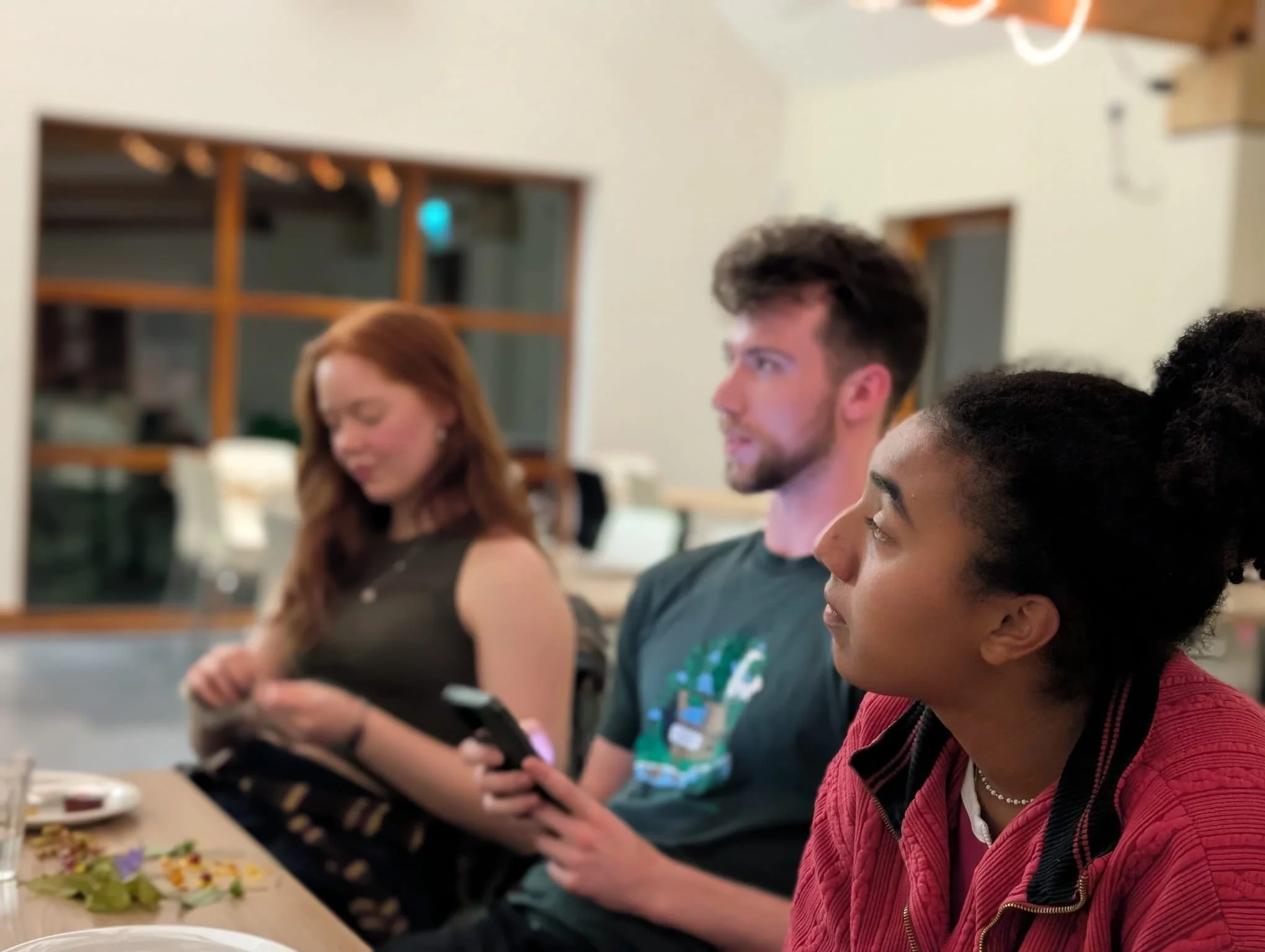 Three young adults seated at a table during a gathering, with food and drinks visible in the foreground. They are engaged and appear to be looking at something off-camera, in a warmly-lit room with wooden framed windows.