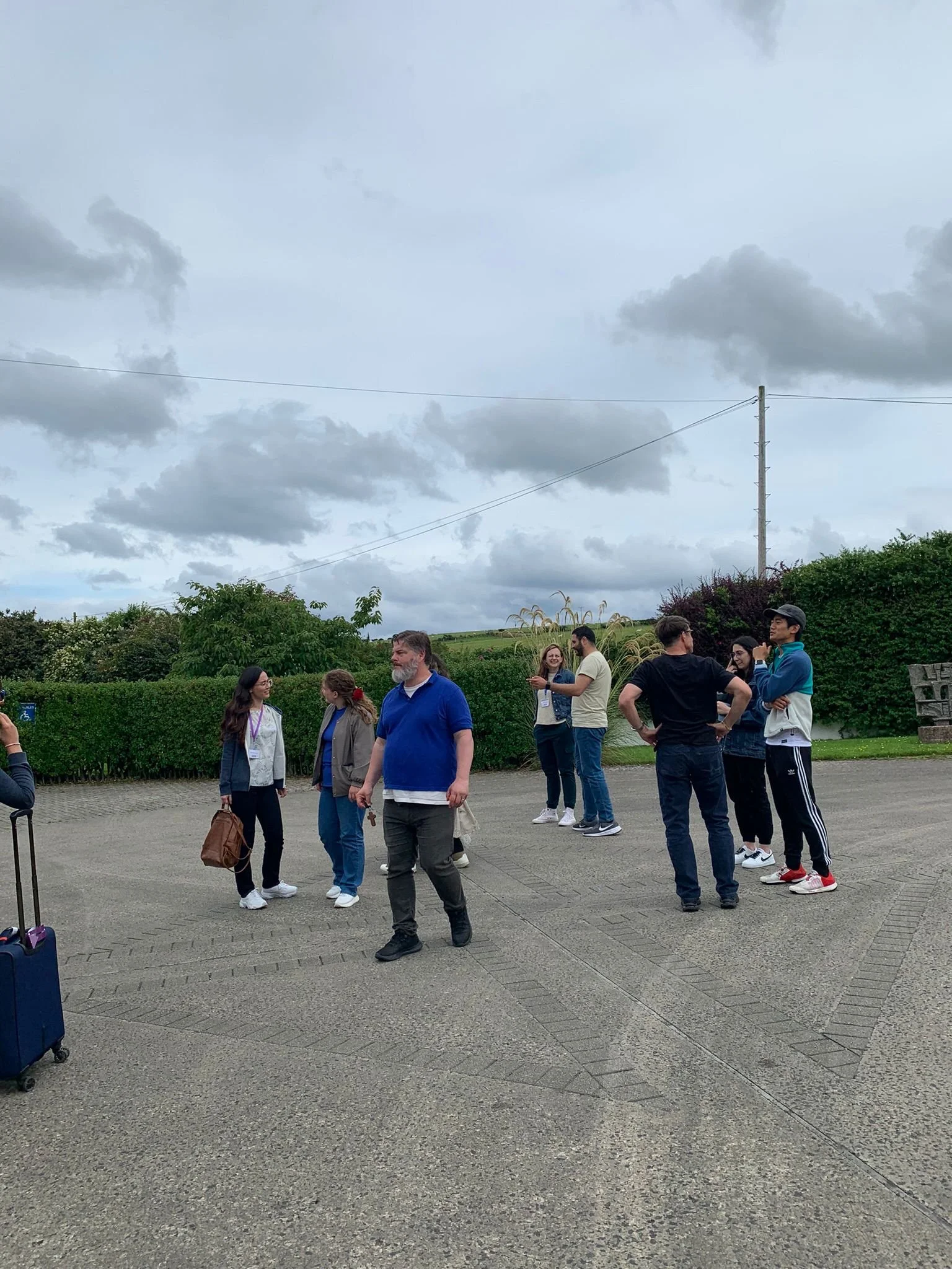 A group of people standing outside on a paved area under a cloudy sky, some with luggage, engaging in conversation.