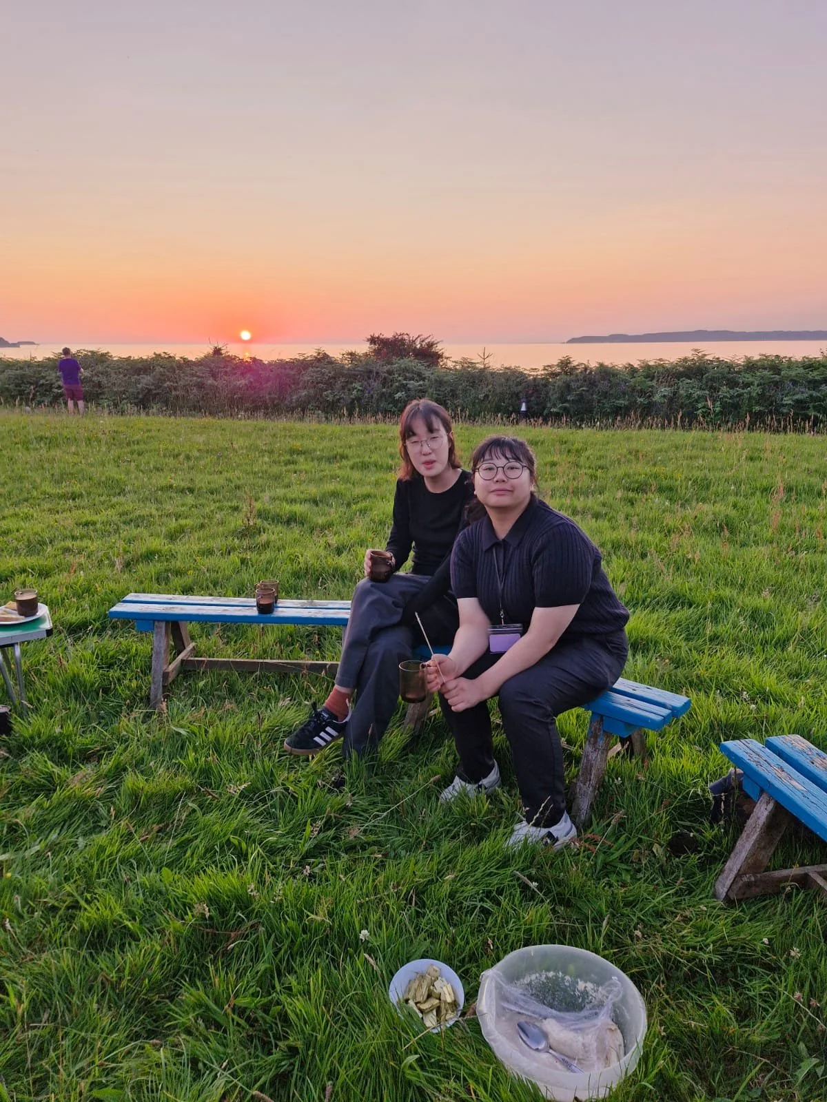Two women sitting on a blue wooden bench on green grass, holding black glasses with a sunset over water in the background. There are plates and food on the ground nearby.