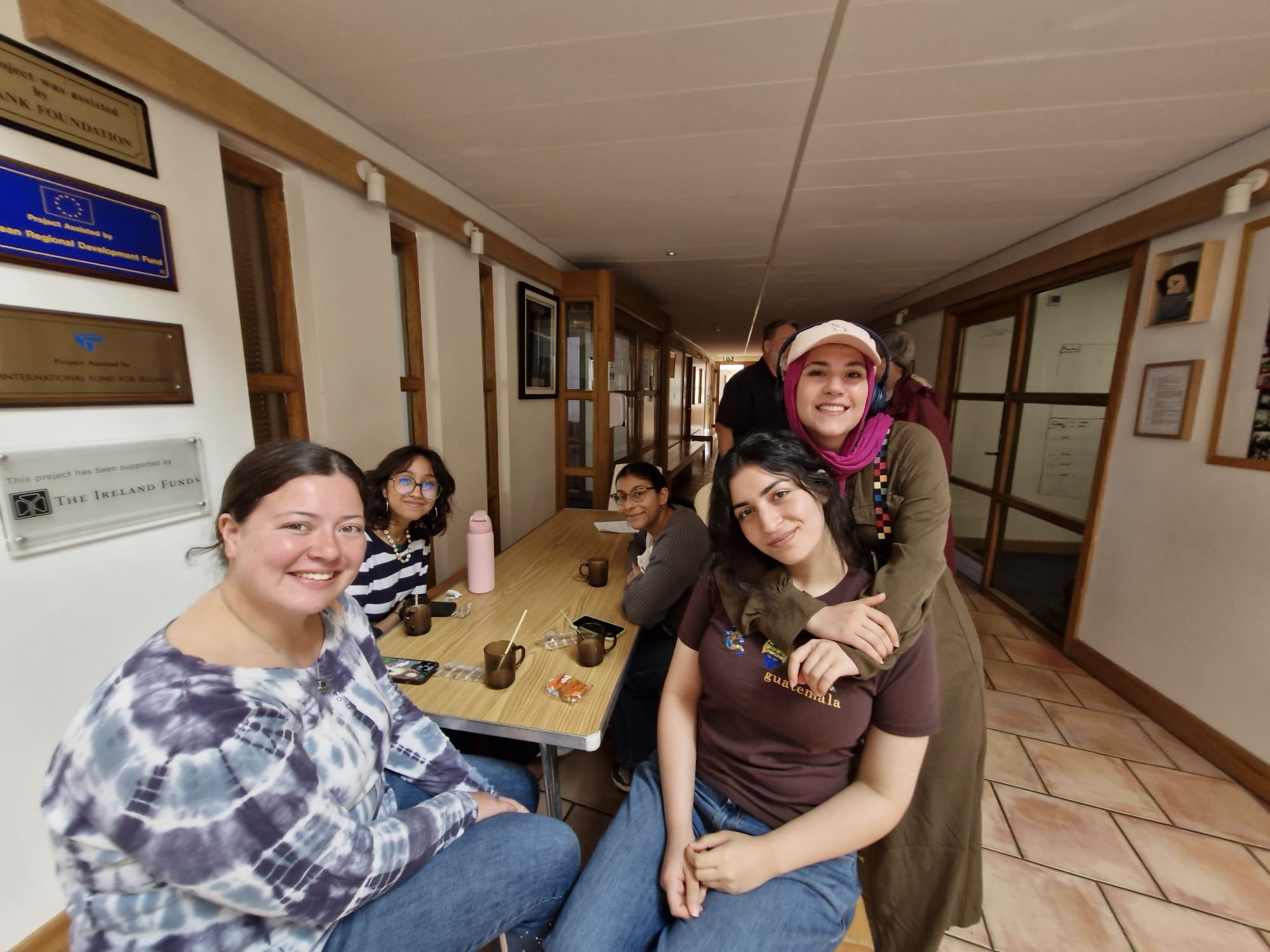 A group of young women sitting around a table, smiling and enjoying a coffee or tea break in a room with certificates and plaques on the walls.