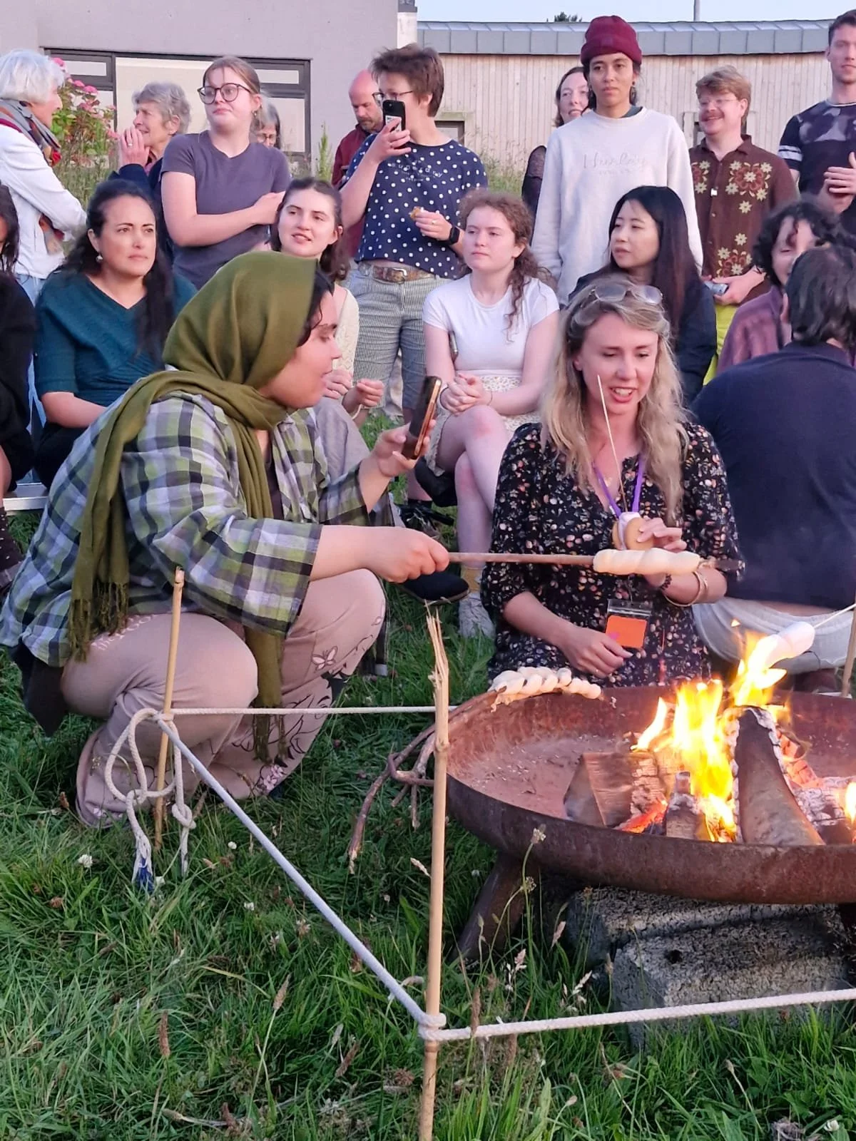 People gathered around a fire pit at an outdoor event, some cooking food on sticks, with a diverse crowd of adults and children observing and engaging in the activity.