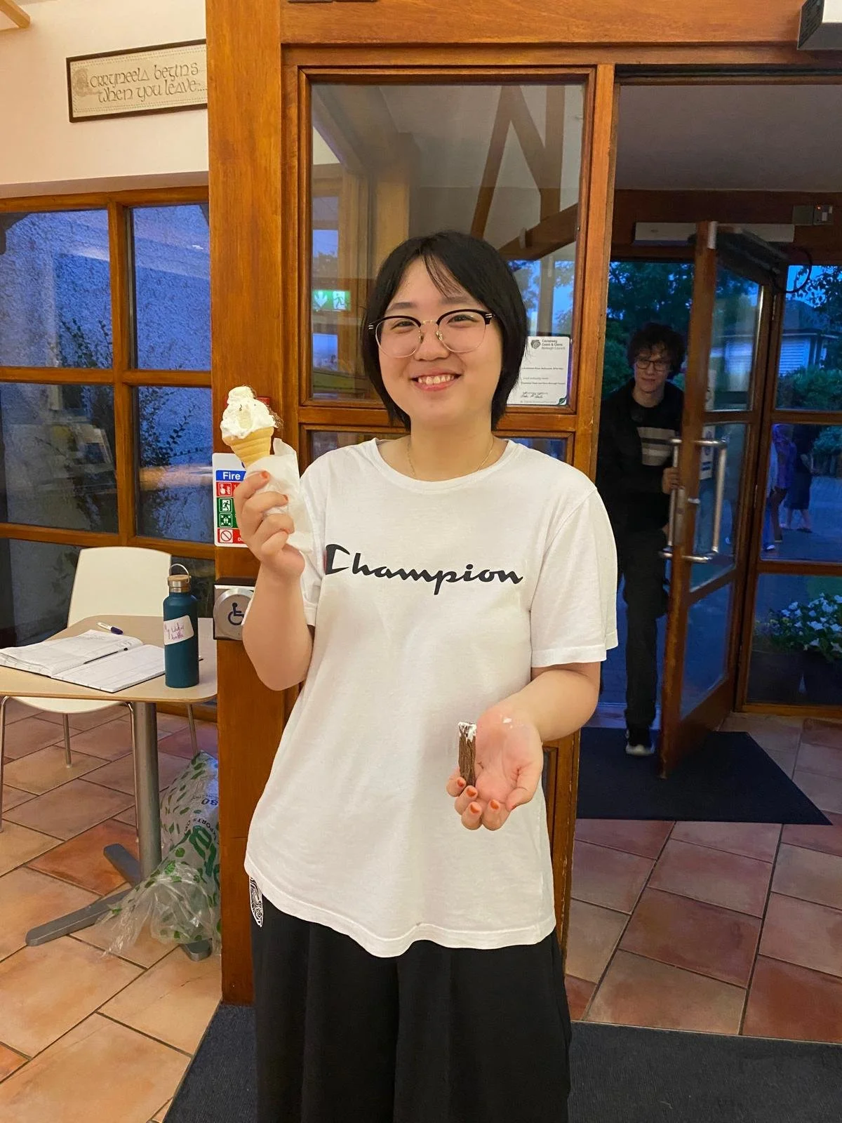 A smiling woman with glasses holding an ice cream cone in one hand and a chocolate treat in the other, inside a restaurant with wooden and glass decor.