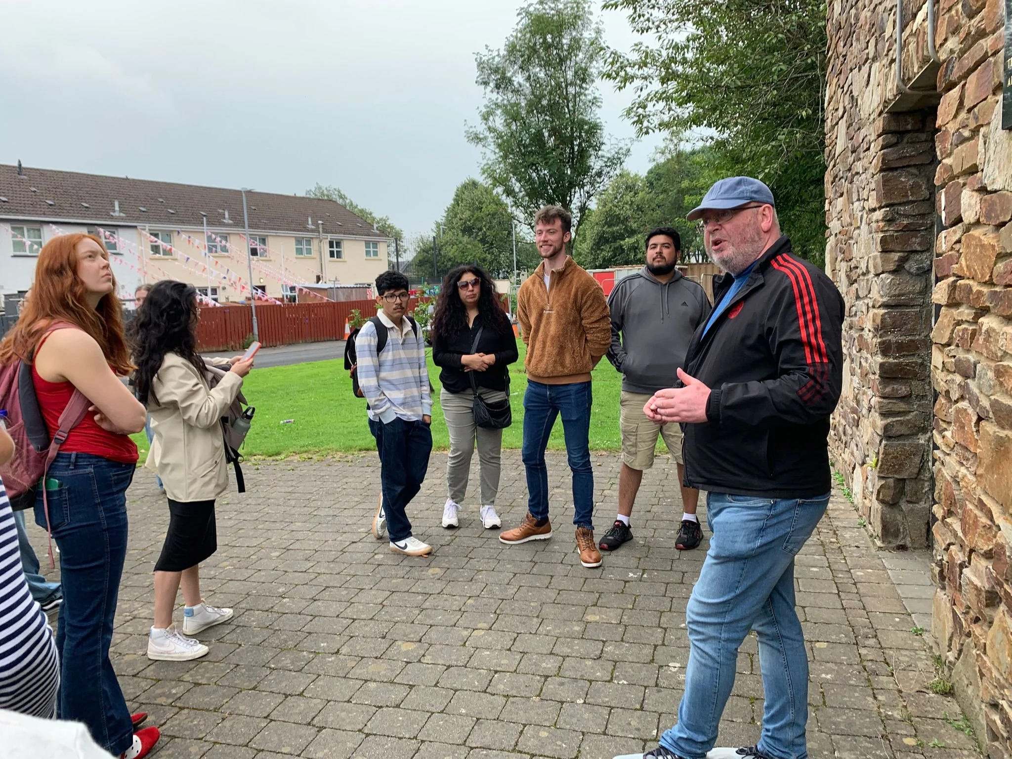 A man with a blue hat speaking to a group of diverse young adults outdoors, next to a stone wall, with houses and greenery in the background.