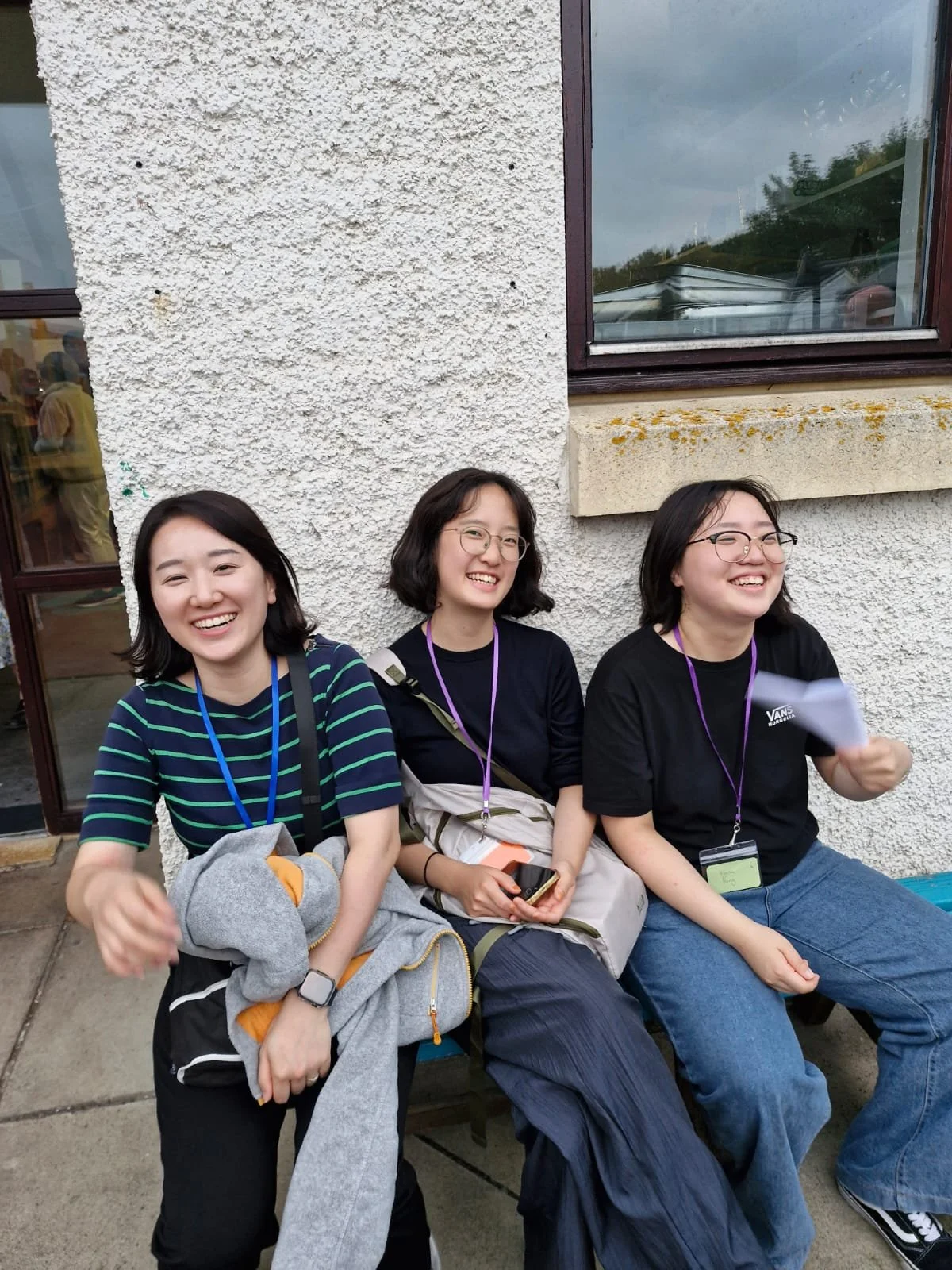 Three young women sitting on a bench outside a building, smiling and laughing, wearing casual clothing and glasses, with backpacks and a jacket.