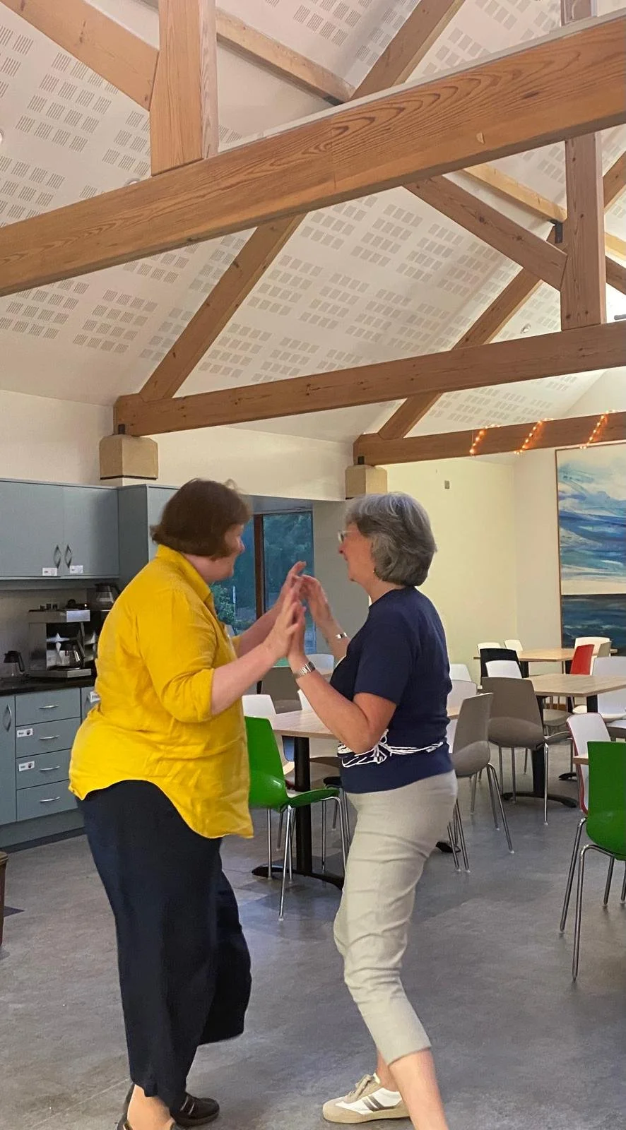 Two women dancing and smiling in a brightly lit room with high wooden beams and modern decor.