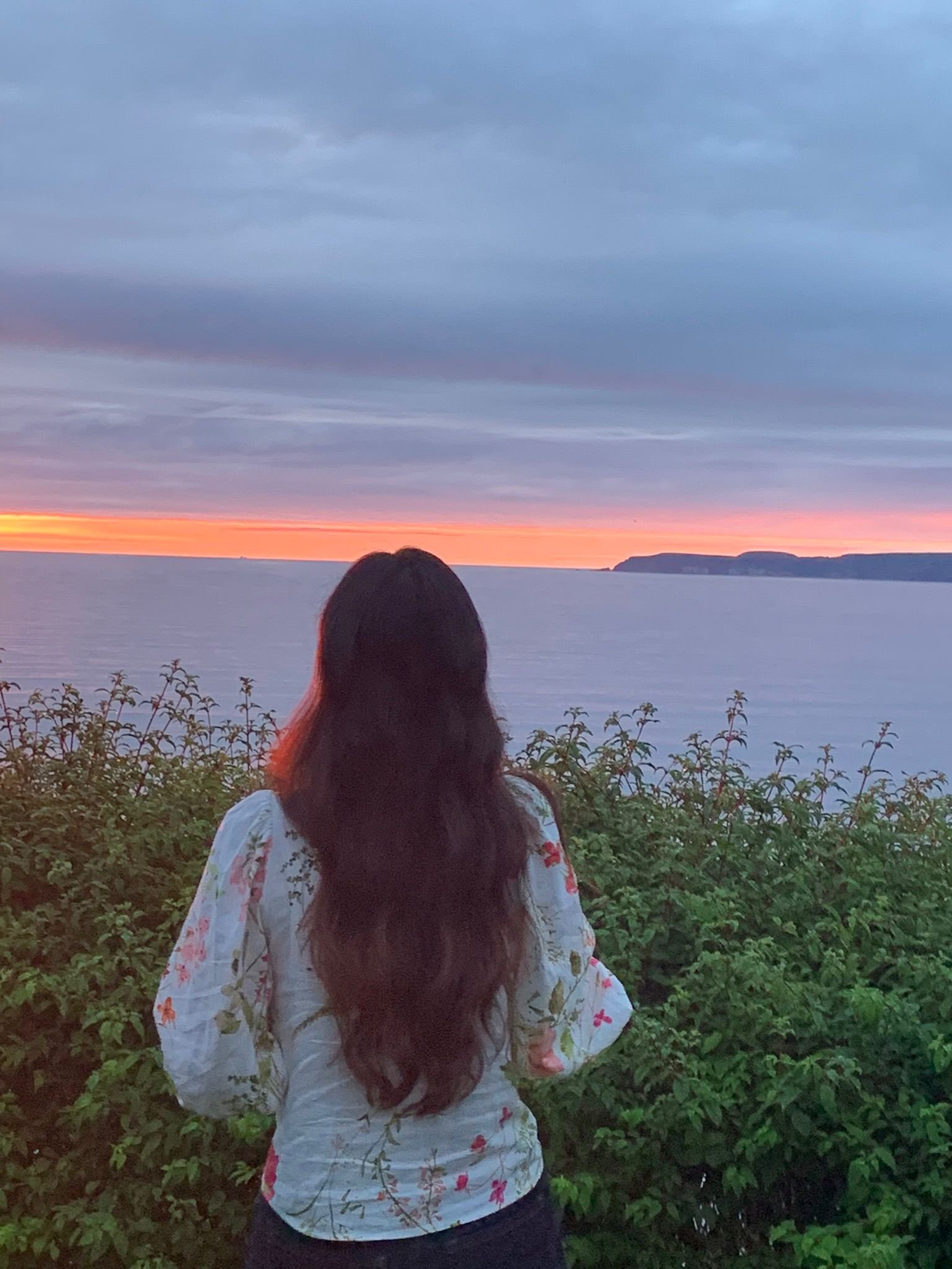 A woman with long brown hair, wearing a white blouse with floral patterns, stands with her back facing the camera, watching a sunset over the ocean with foliage in the foreground and a distant landmass on the horizon.
