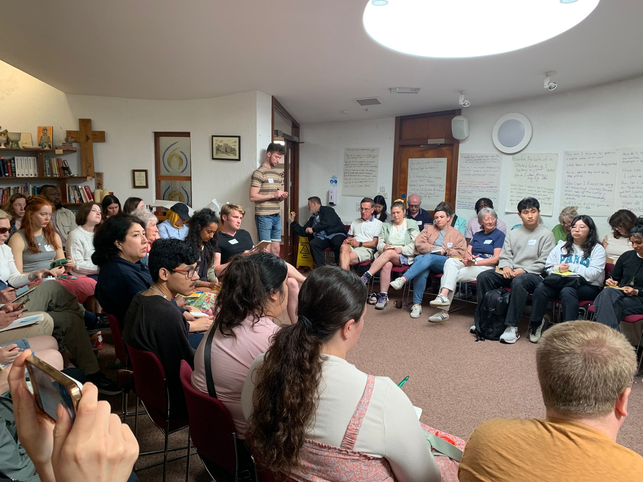 Group of people seated in a circle in a room, engaging in a discussion or workshop, with some taking notes.