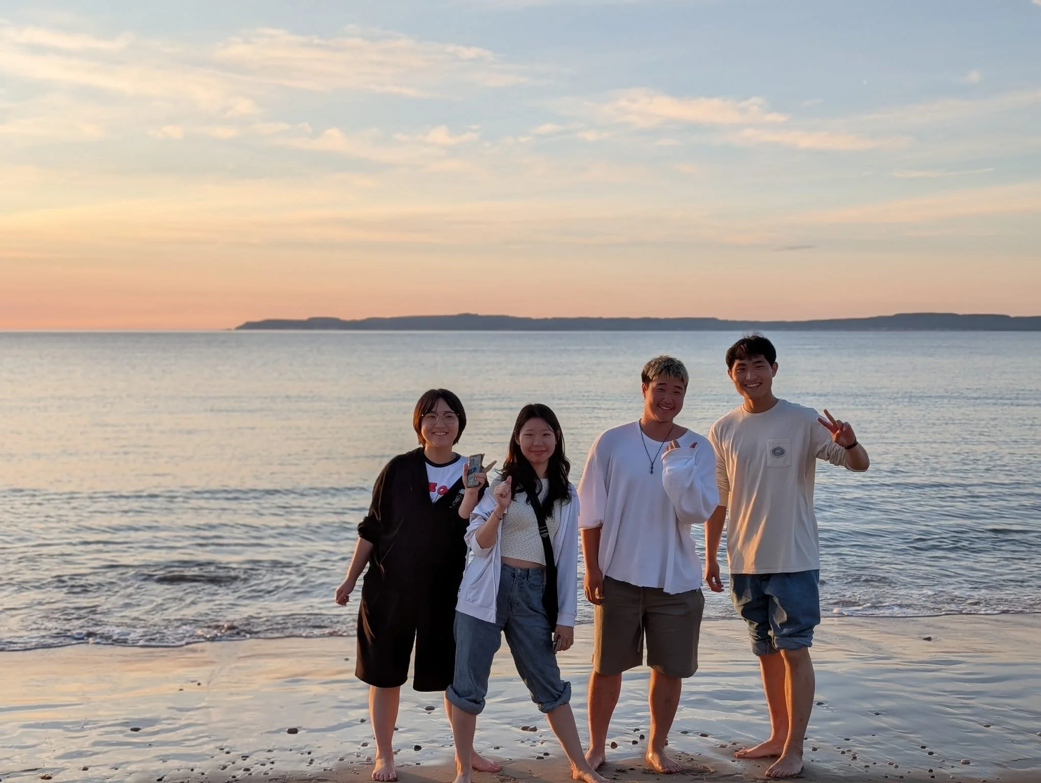 Four young adults standing on a beach during sunset, smiling and posing for a photo.