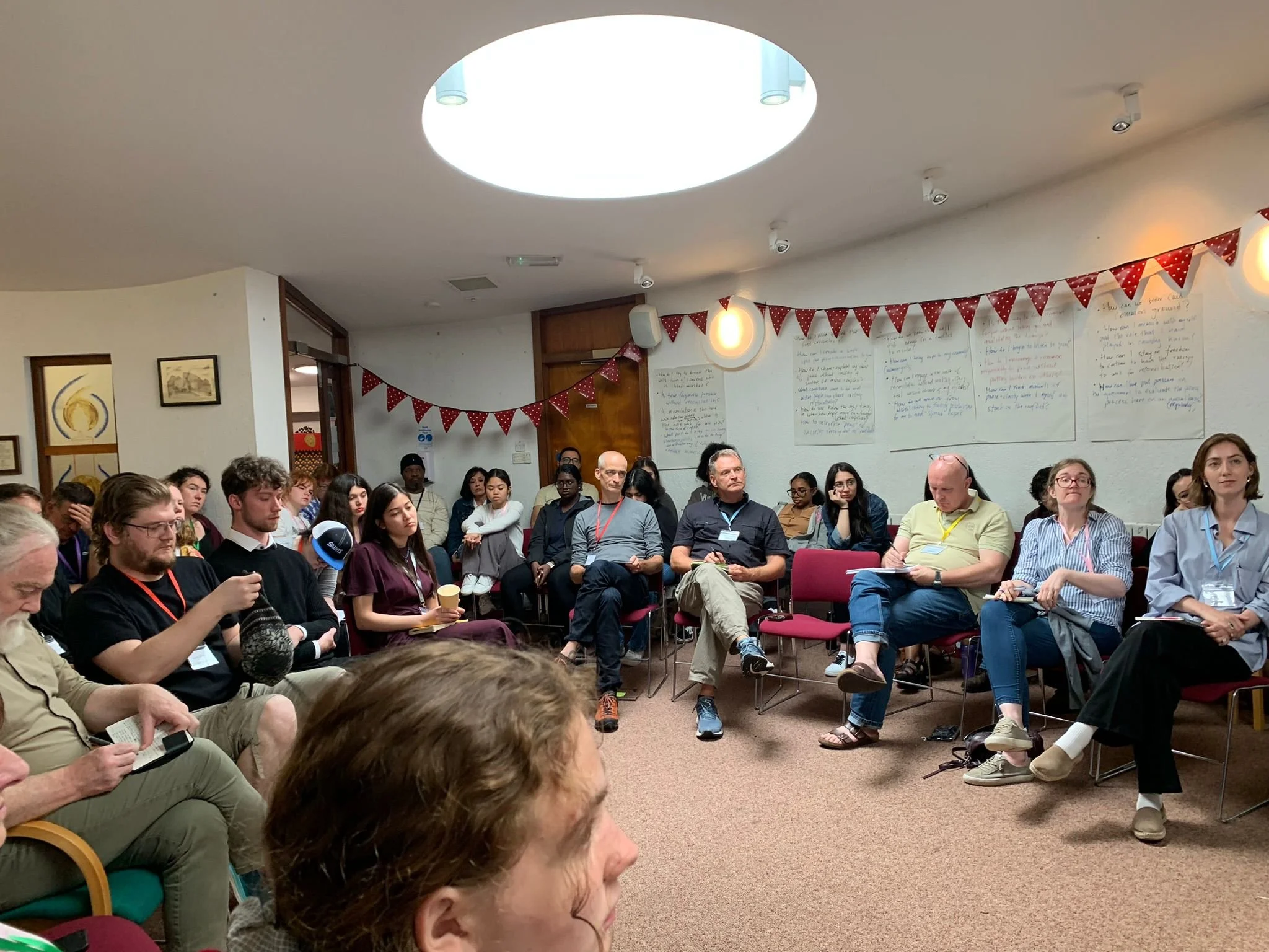 People seated in chairs attending a discussion or presentation in a conference room, with some taking notes, listening, or looking at their devices; room decorated with bunting and wall writings.