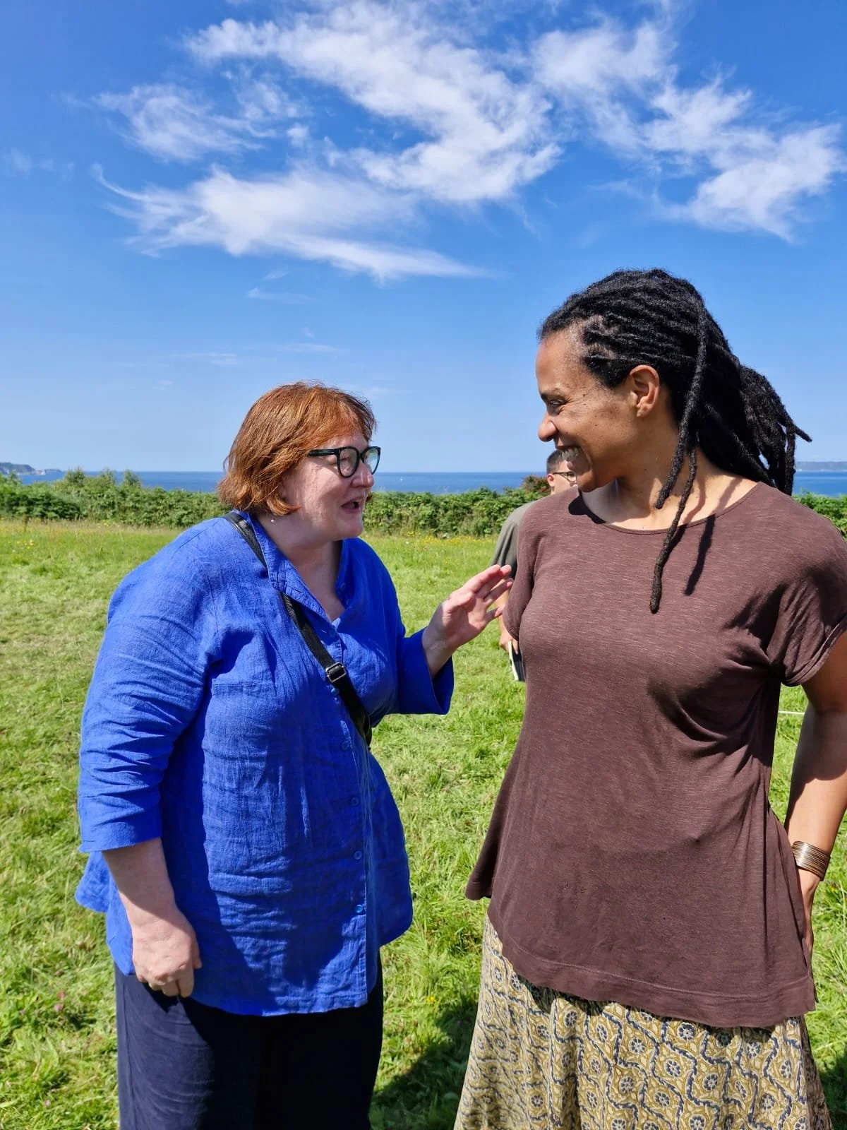 Two women talking and smiling outdoors in a grassy field with a view of the ocean and blue sky with clouds.