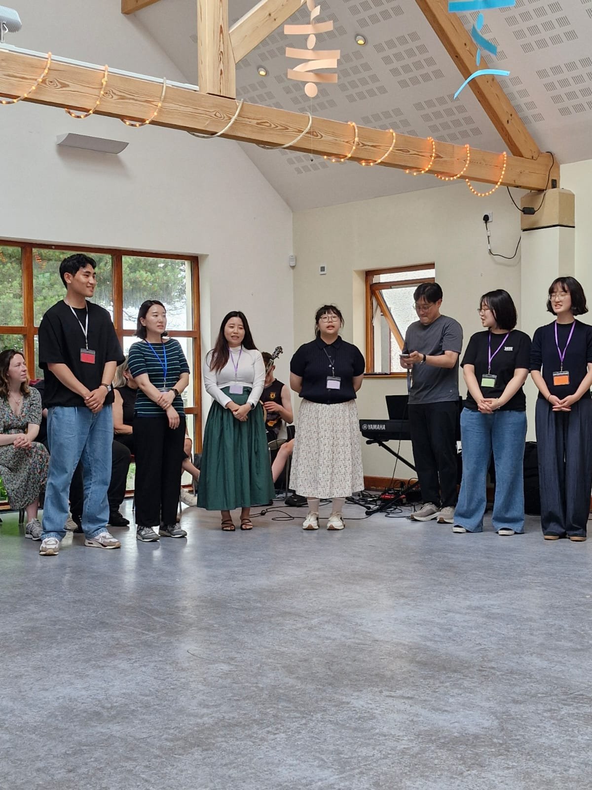 Group of people standing in a circle, with some holding microphones, in a room with wooden beams and large windows, possibly during a performance or presentation.