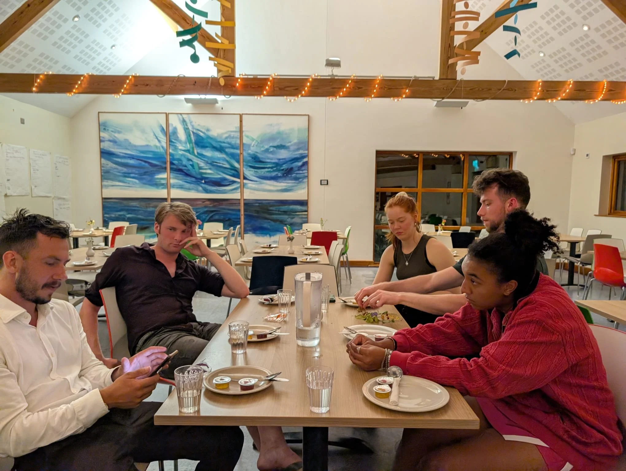 Group of five young adults sitting around a dining table in a restaurant. Three are engaged with their phones or in thought, while two are reaching for items on the table. The restaurant has colorful chairs, a large water-themed painting on the wall,