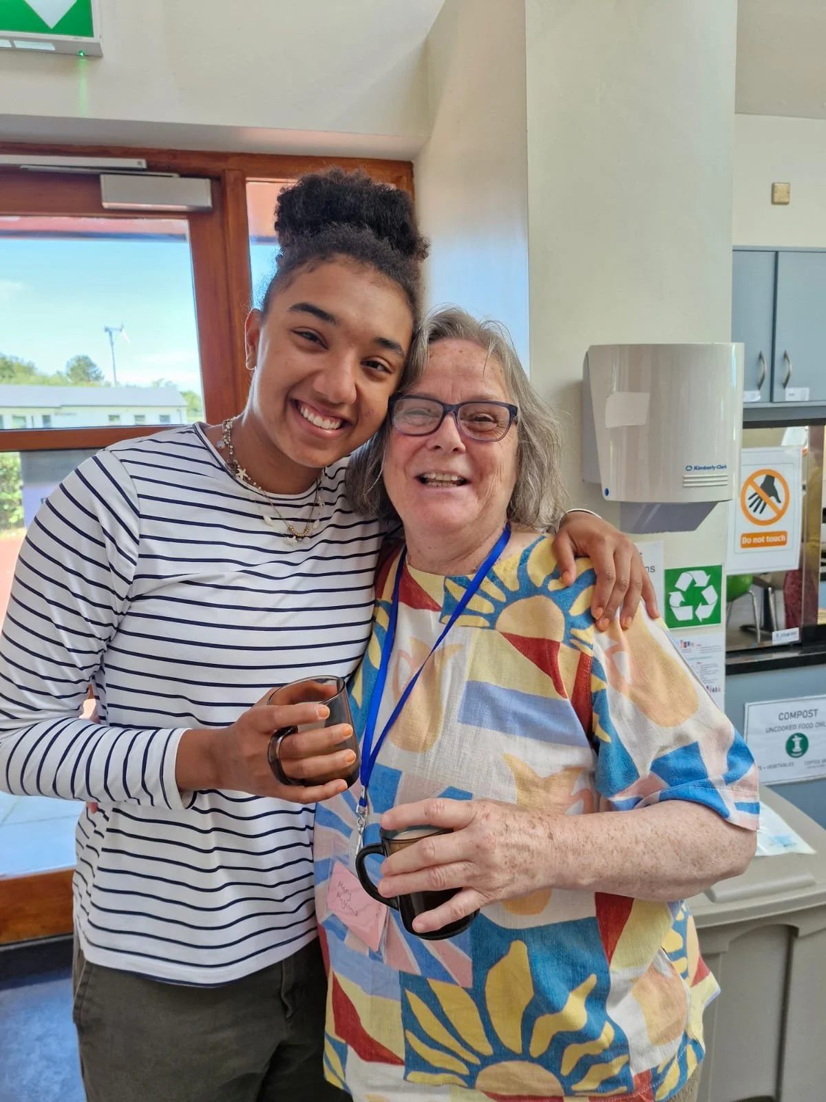 Two women smiling and hugging each other in a room; one younger woman with dark, curly hair in a bun, wearing a striped shirt, and an older woman with glasses, gray hair, and patterned shirt, holding a black mug.