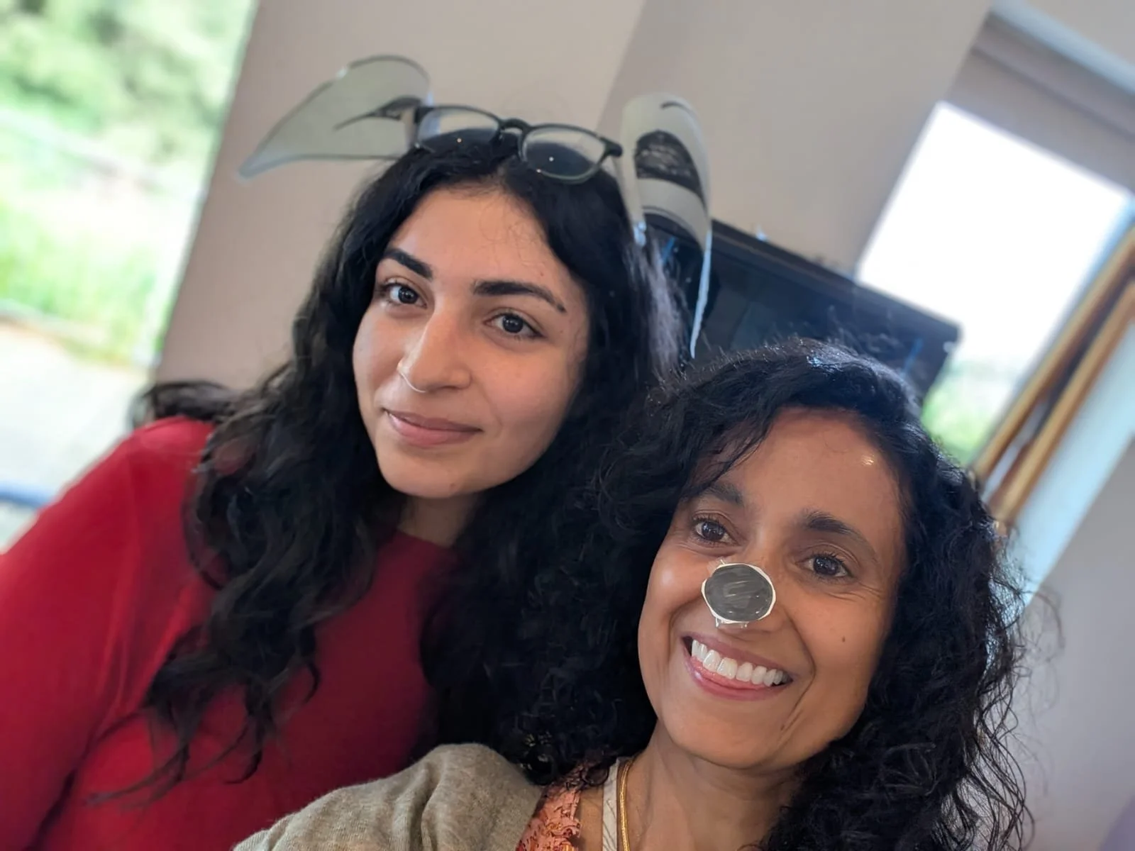 Two women with curly hair smiling close together indoors, with busts of bald men wearing glasses on their heads in the background.