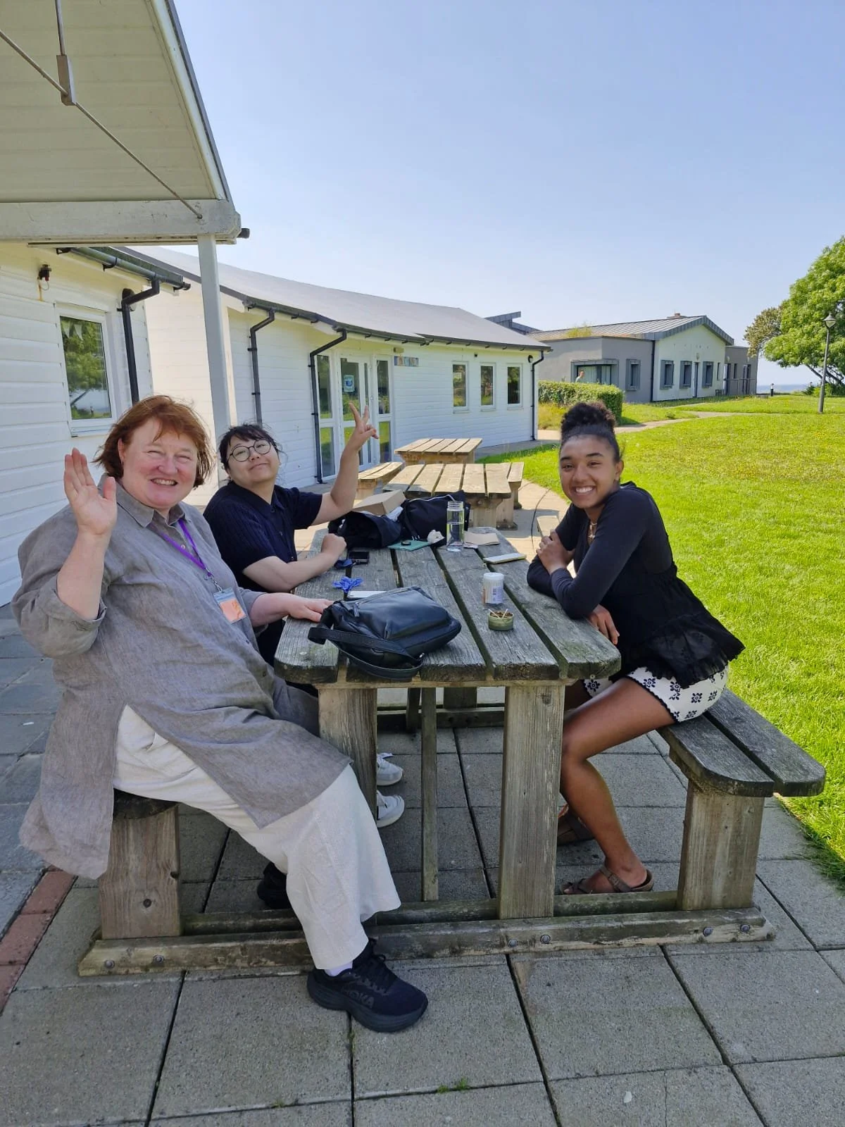 Three women sitting at an outdoor wooden picnic table with a white building and green lawn in the background. They are smiling and waving, with various items like bags and papers on the table.