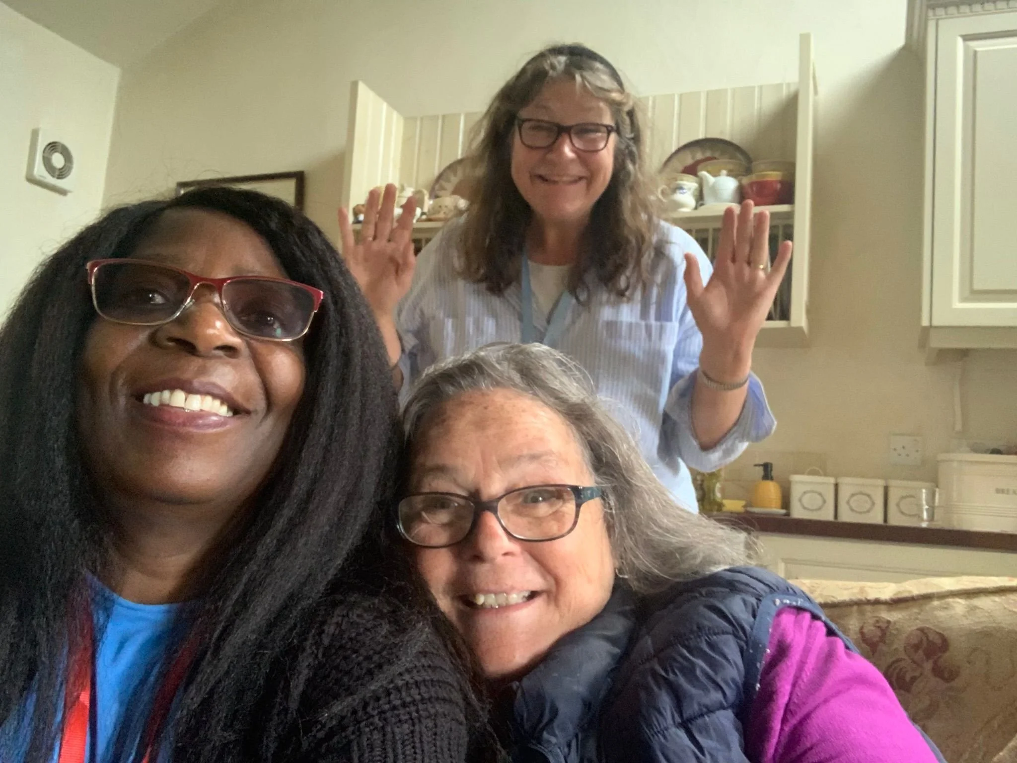 Four women gathering together indoors, smiling and posing for a selfie. One woman in the background has her hands raised with a cheerful expression.