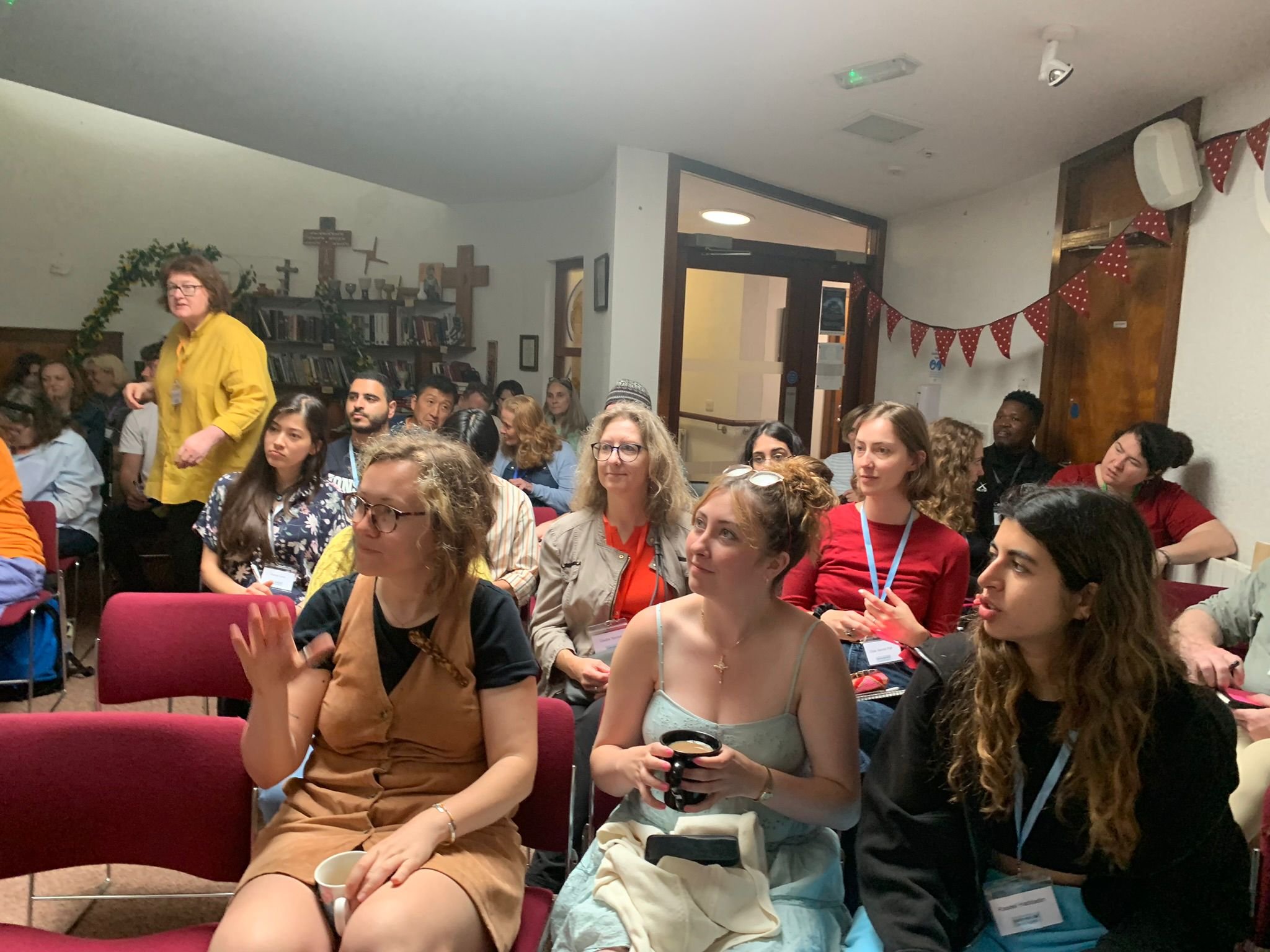 Group of people seated in an indoor conference or seminar, listening attentively, with some taking notes, wall decorations, and a woman in a yellow shirt standing at the front.