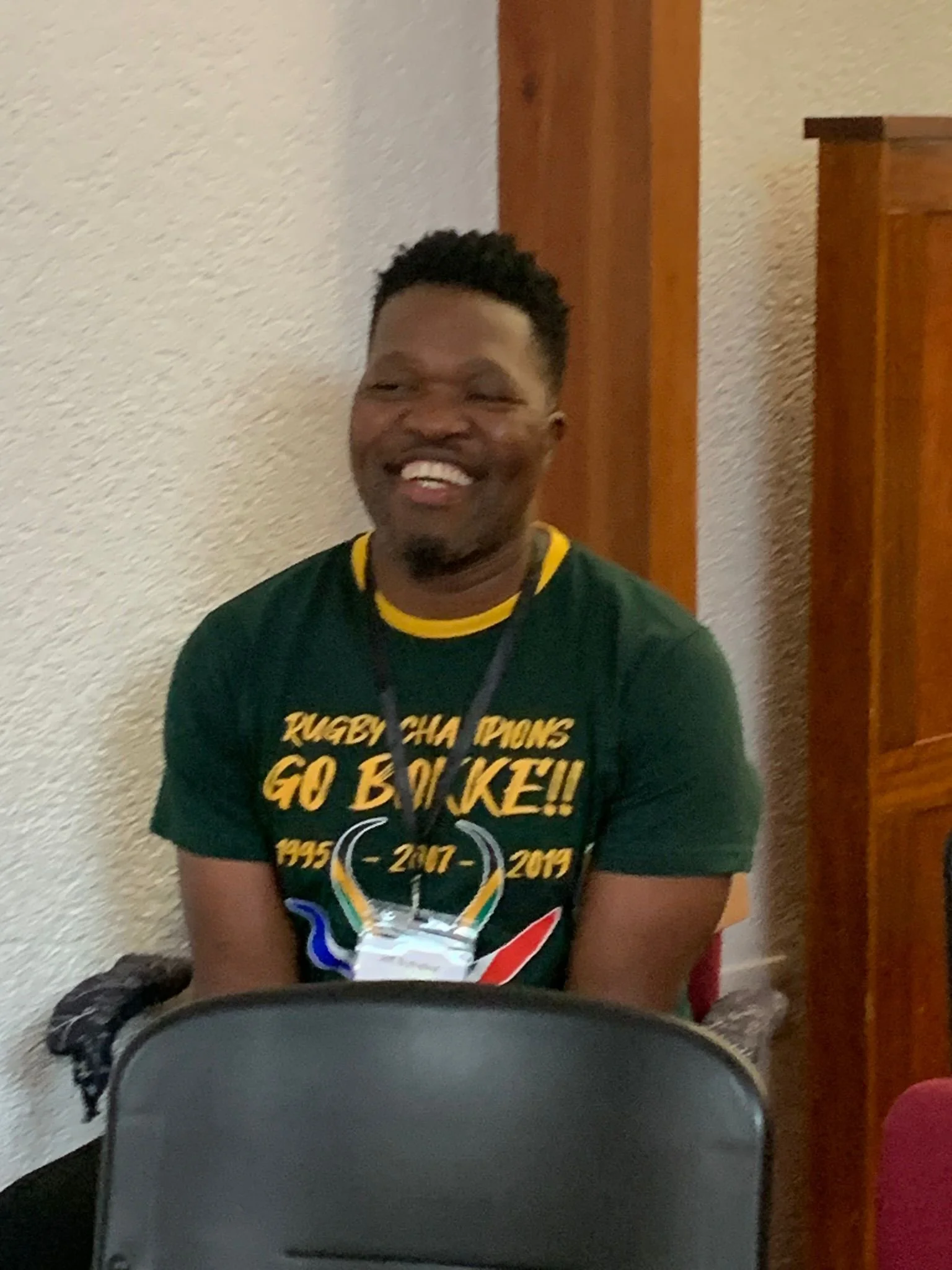 A man with dark hair and a beard smiling while sitting in a room with beige walls and wooden furniture, wearing a green T-shirt that has yellow and red text and graphics related to a rugby league championship.