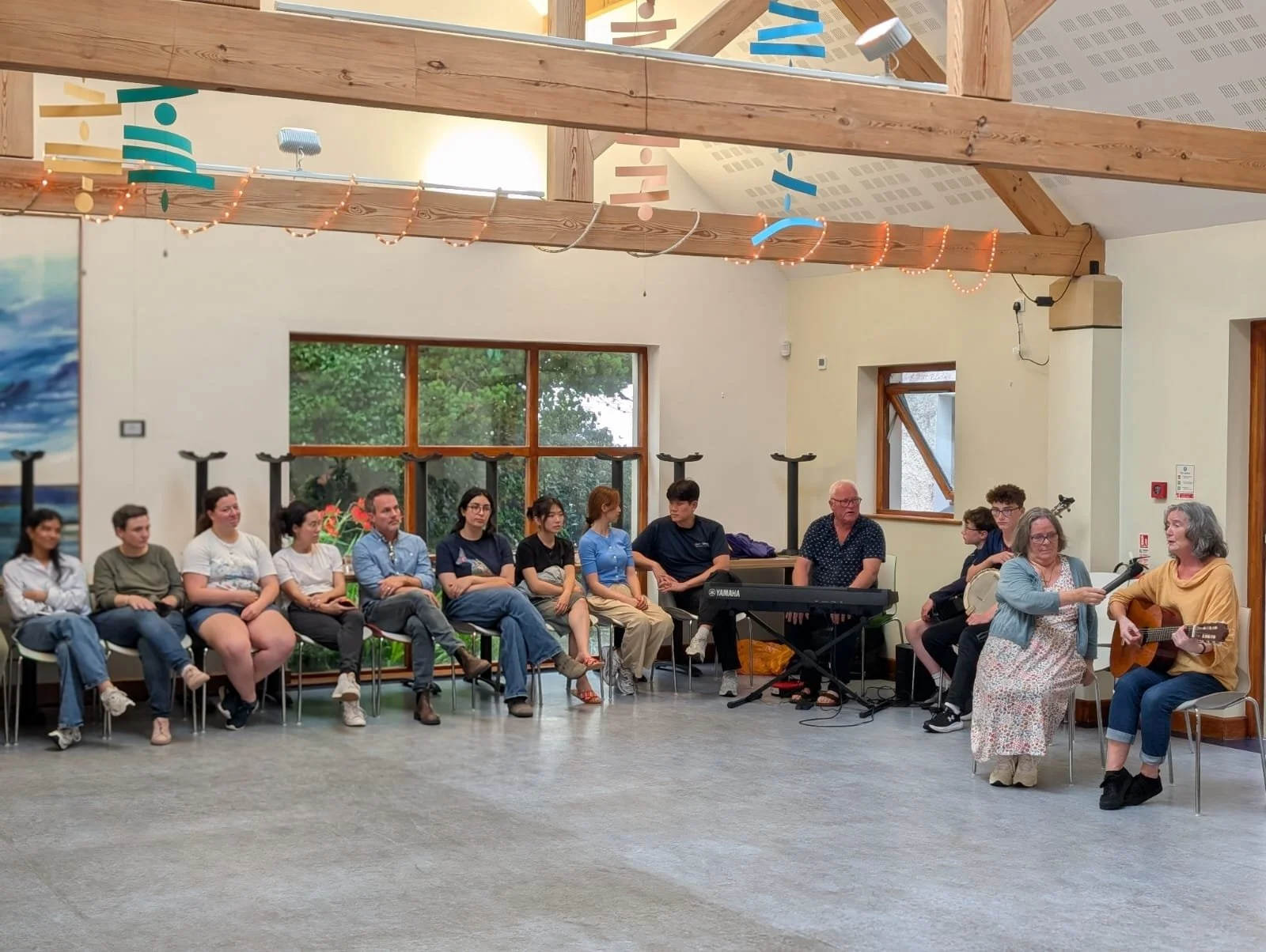 A group of people sitting inside a room with wooden beams, large windows, and colorful hanging decorations, with two women at the front playing guitar and singing.