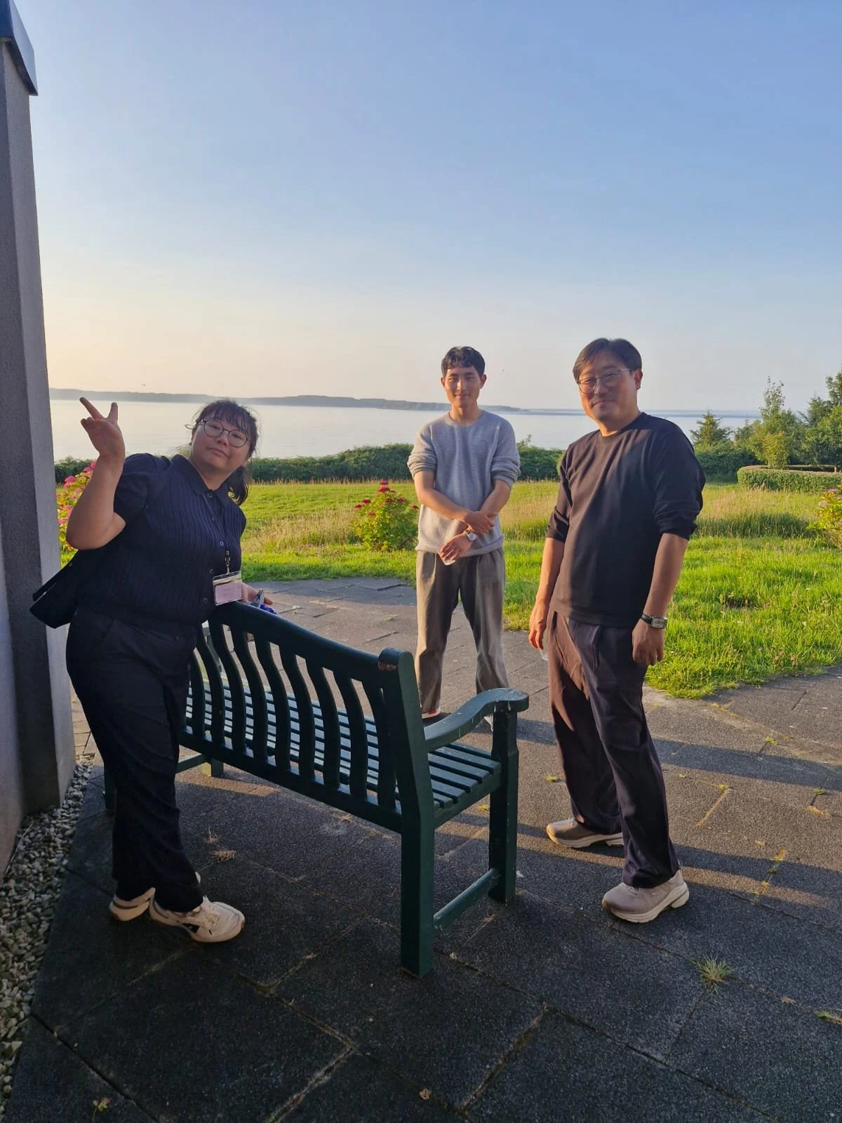 Three people standing outdoors on a paved area near a grassy field with trees and a body of water in the background, during sunset. One woman is on the left making a peace sign, and two men are on the right smiling at the camera.