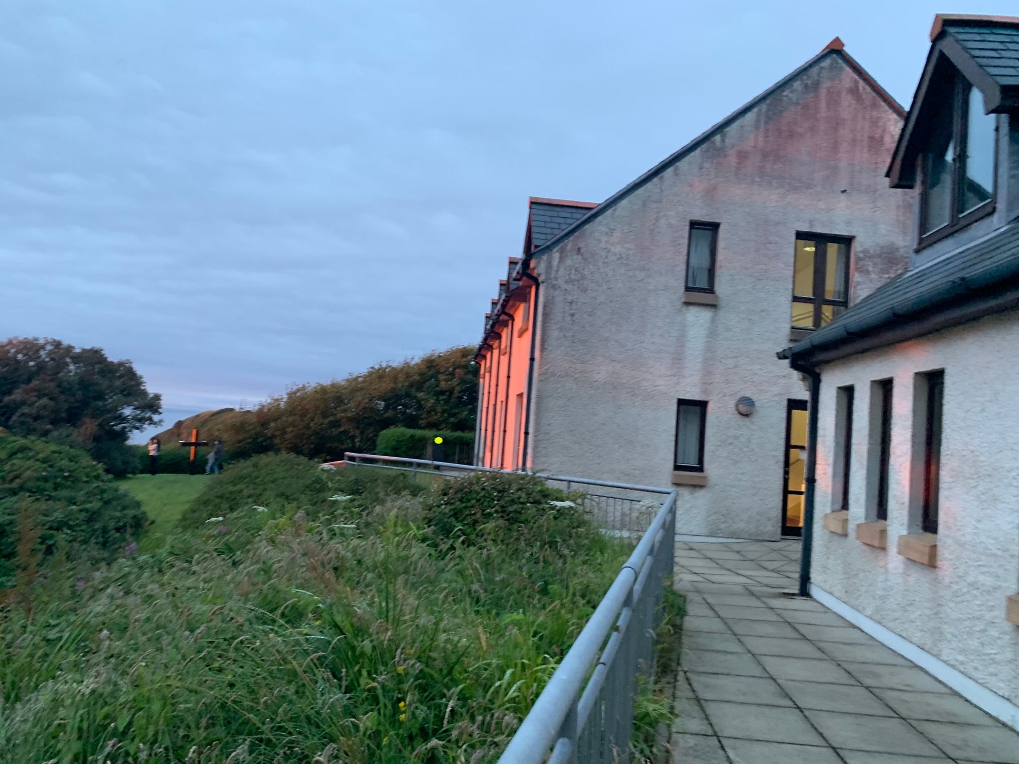 A pathway along a building's exterior with windows, a grassy hillside, trees, and cloudy sky at dusk.