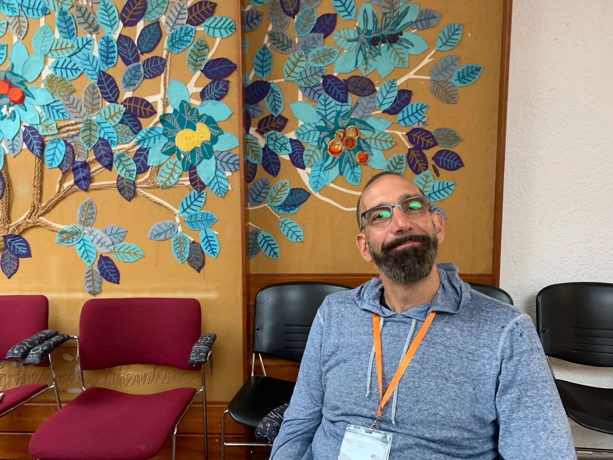 A bearded man wearing glasses, a blue hoodie, and an orange lanyard with a name tag sitting in front of a colorful mural depicting blue and purple leaves and branches on a beige background.