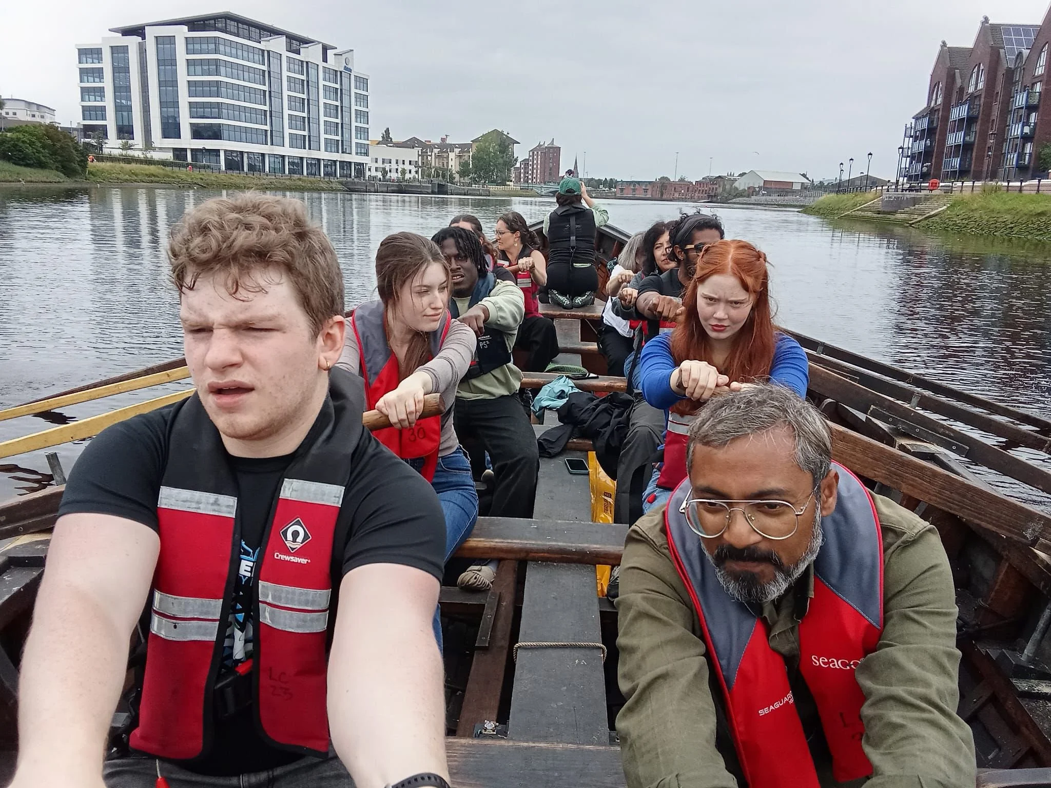 A group of people on a boat paddling on a river. The background shows modern and older buildings along the riverbank.