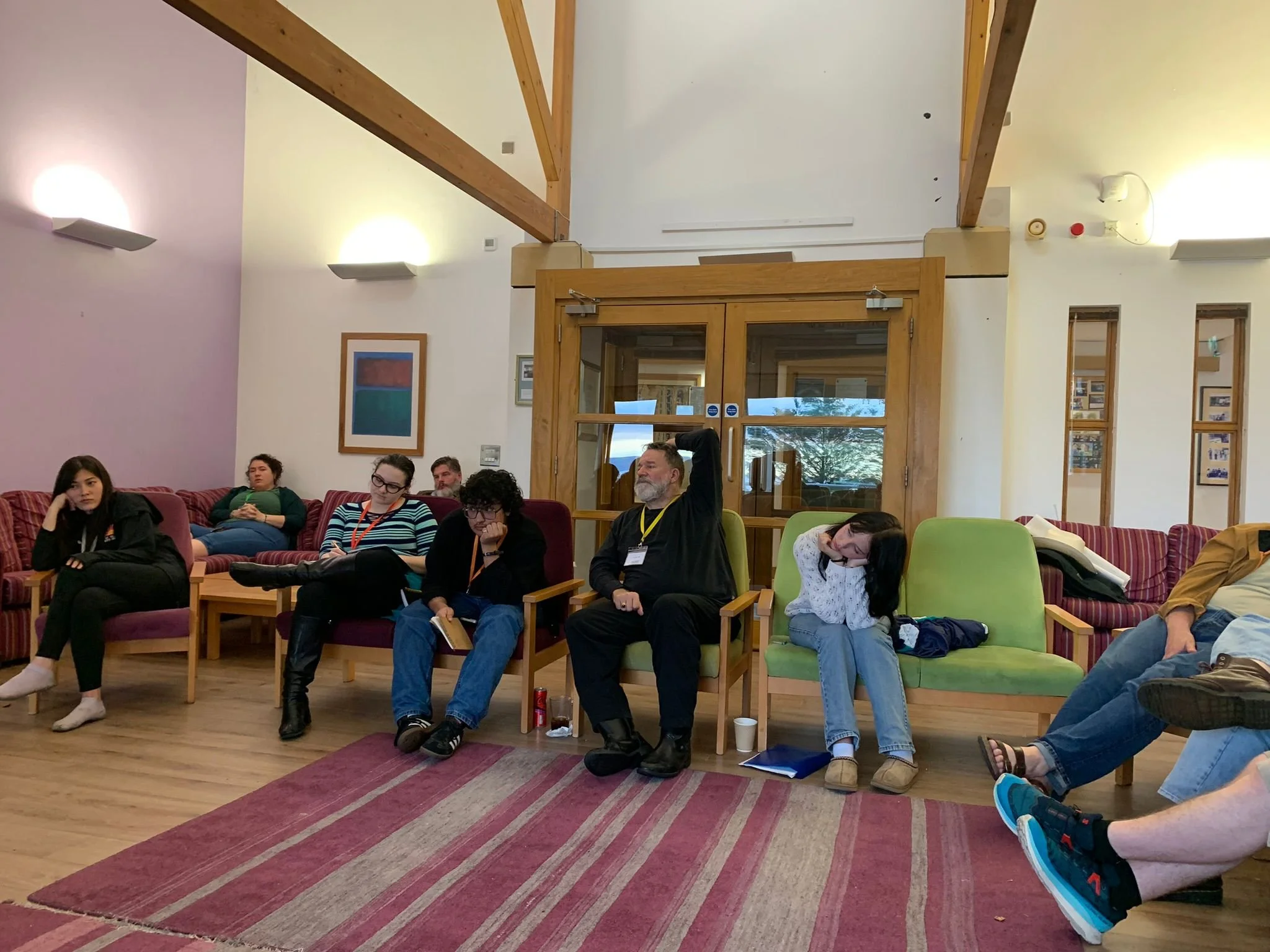 Group of people sitting on chairs and couches in a lounge or meeting room, listening to a presentation or discussion.