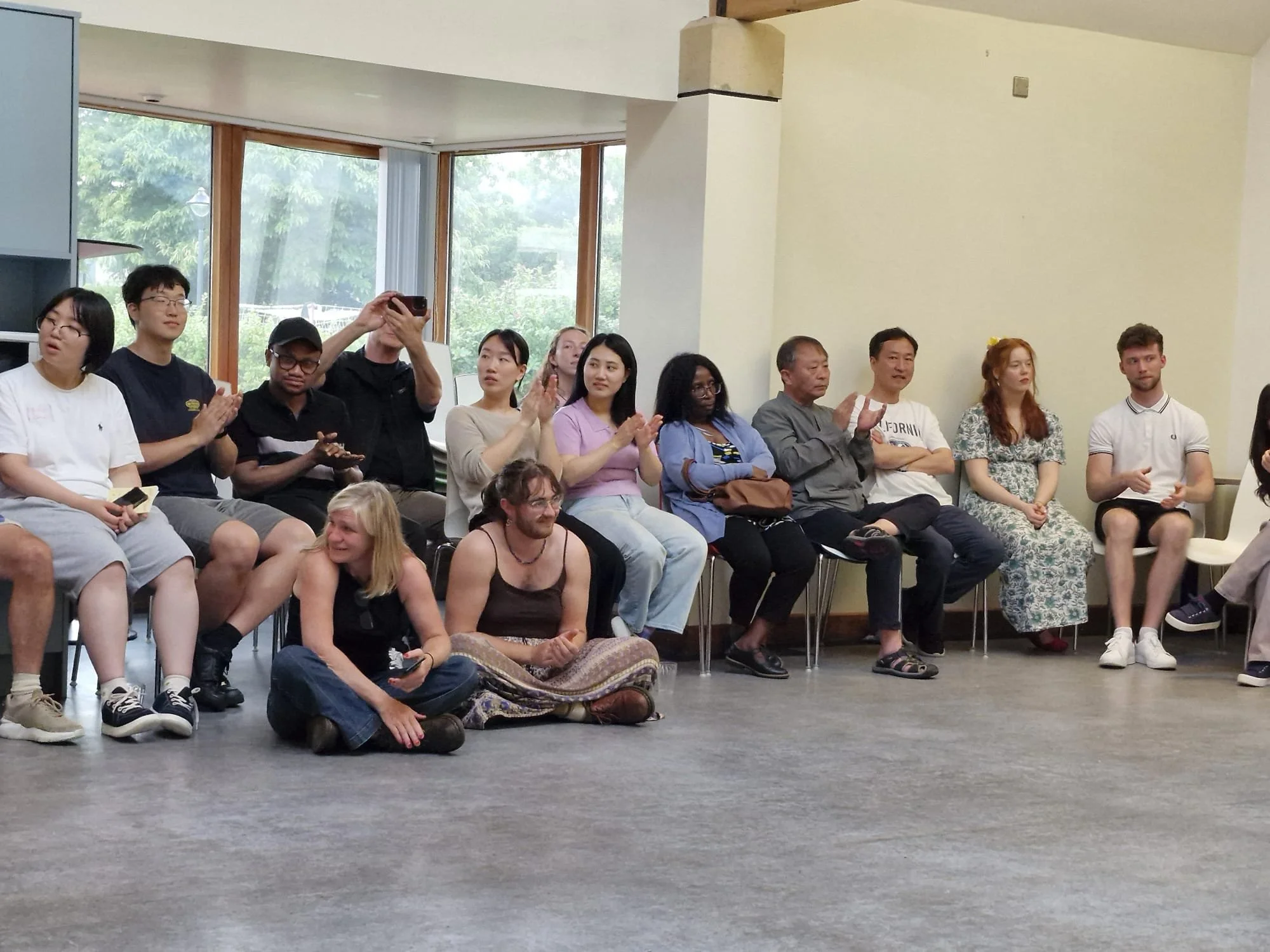 A diverse group of people sitting in a semi-circle, some clapping and one taking a photo, in a room with large windows showing greenery outside.