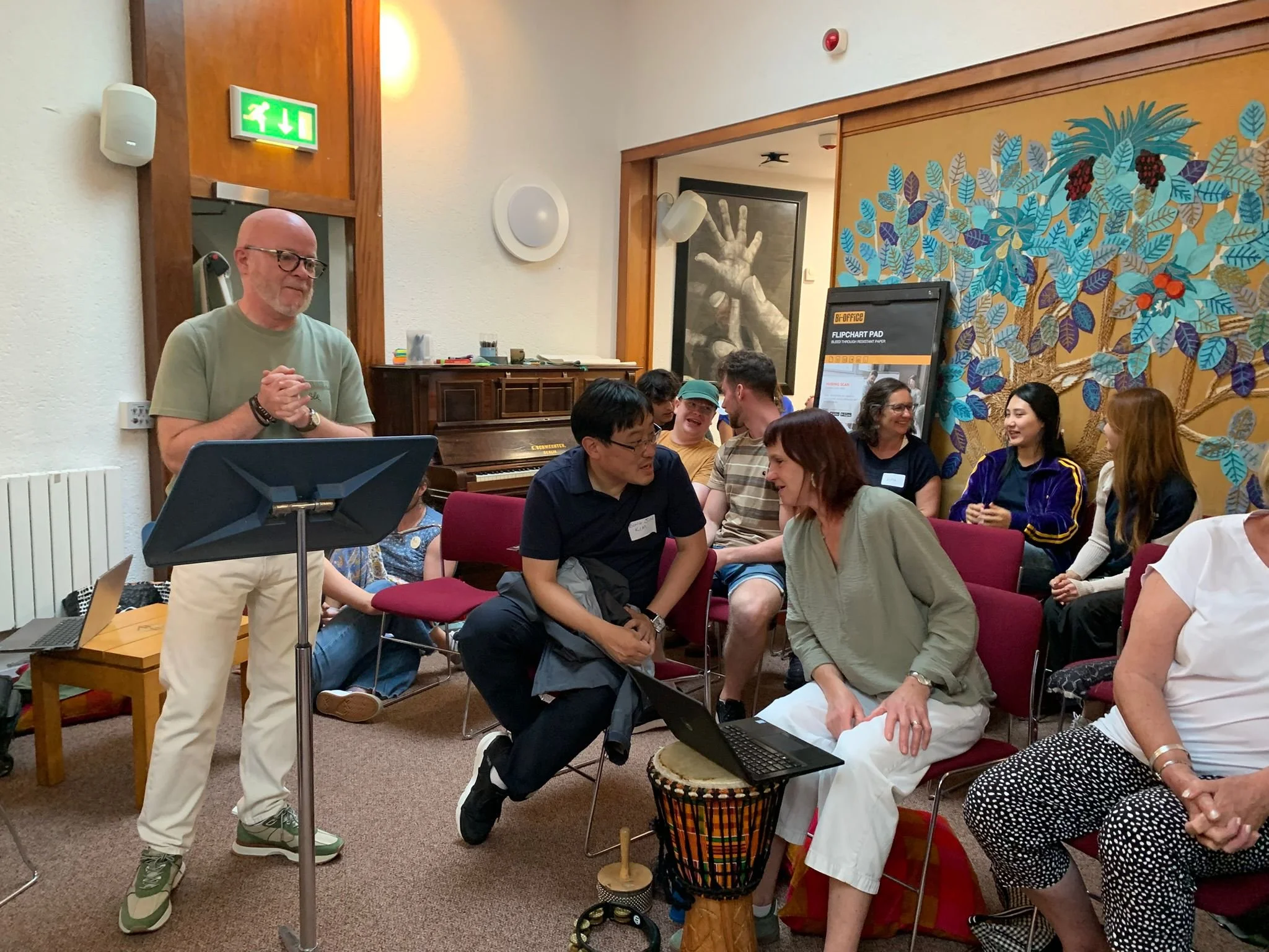 A group of people sitting in chairs in a room, engaging in conversation and smiling, while a man stands at a music stand, possibly leading a discussion or presentation.