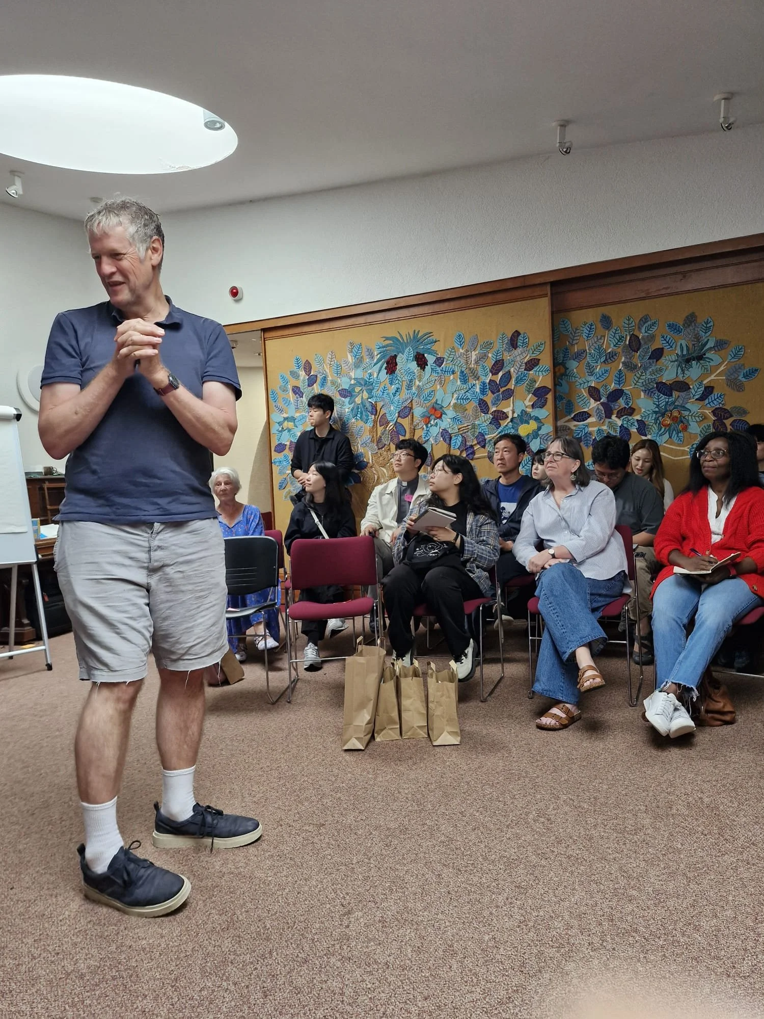 A man in a blue shirt and shorts standing in front of a seated audience in a meeting room with a decorative mural on the wall.