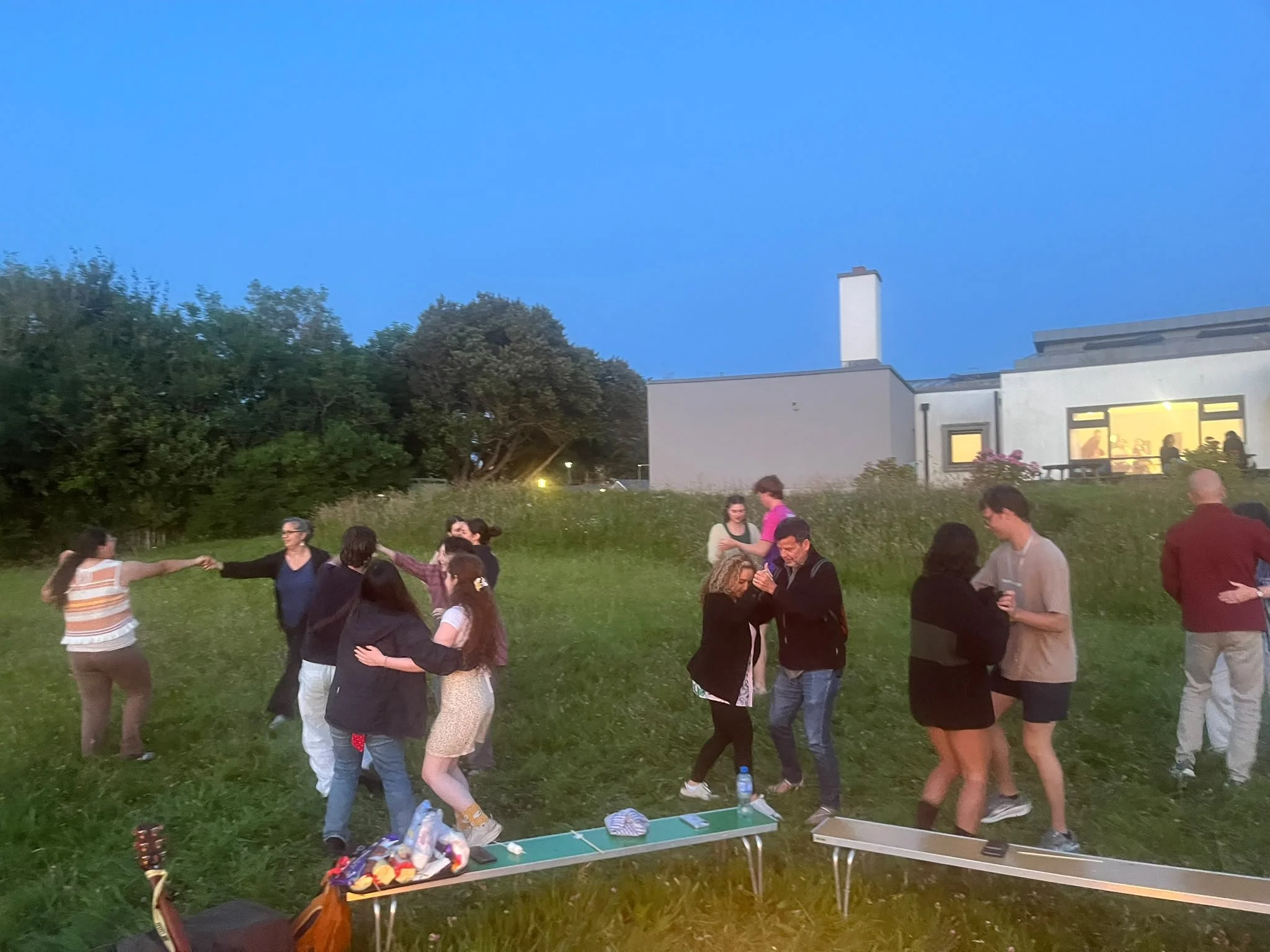 People dancing in a circle outdoors during the evening, with a modern house and trees in the background.