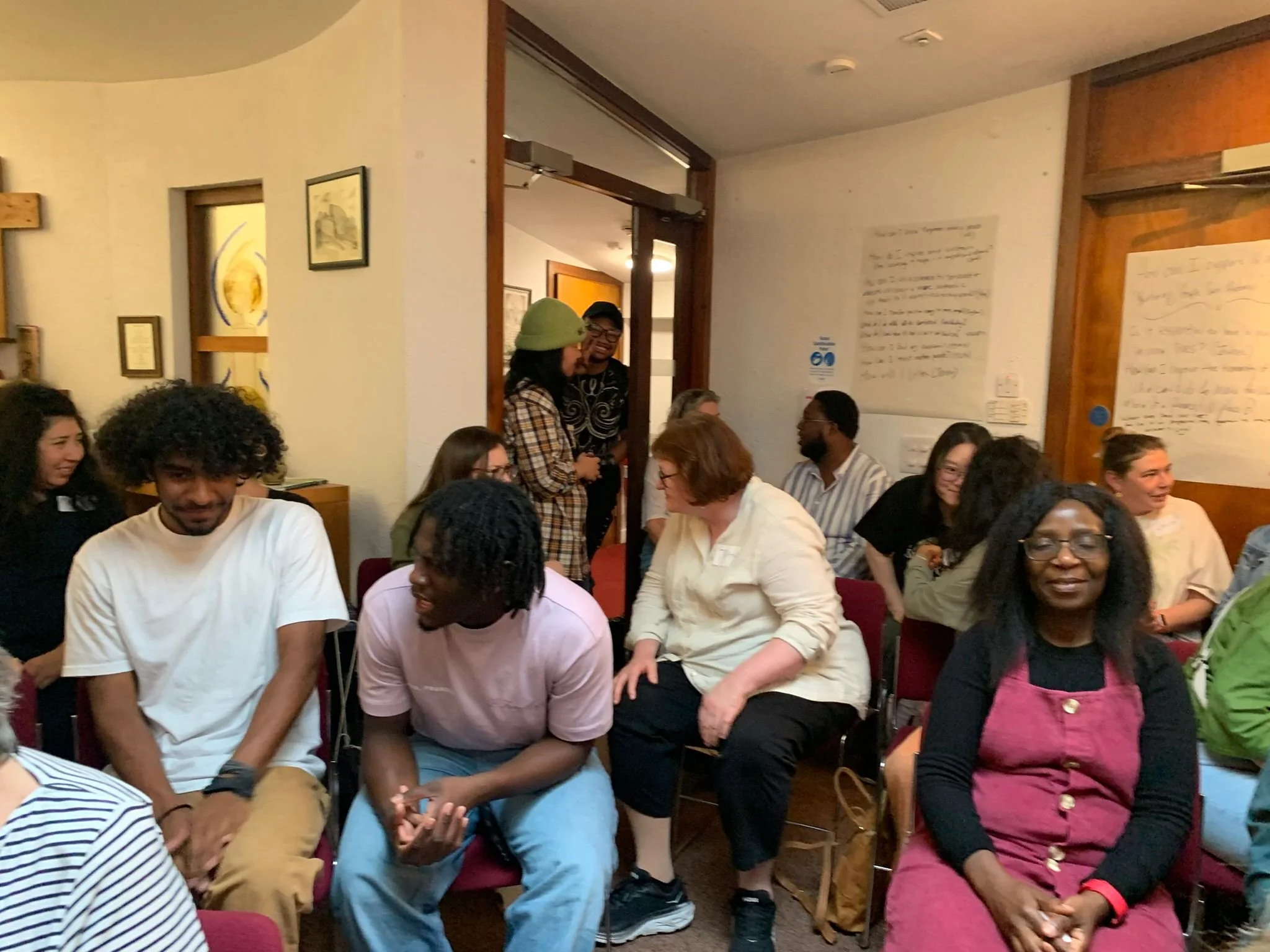 A group of people gathered indoors, some sitting and some standing, engaging in conversations and smiling at each other.