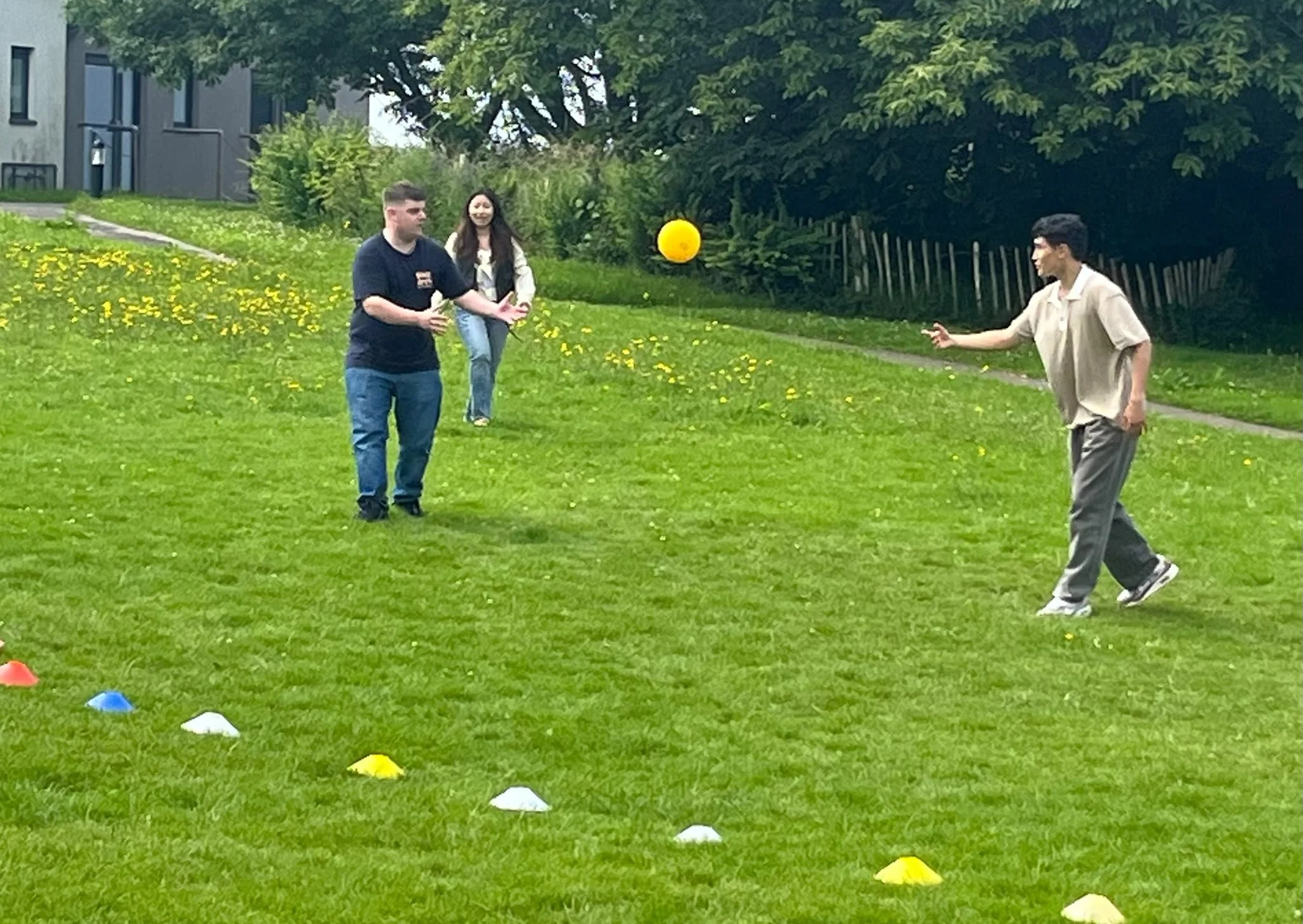 Three people playing a game with a yellow ball outdoors on a grassy area, with one person throwing the ball to another, and a third person smiling in the background, along with trees and buildings.