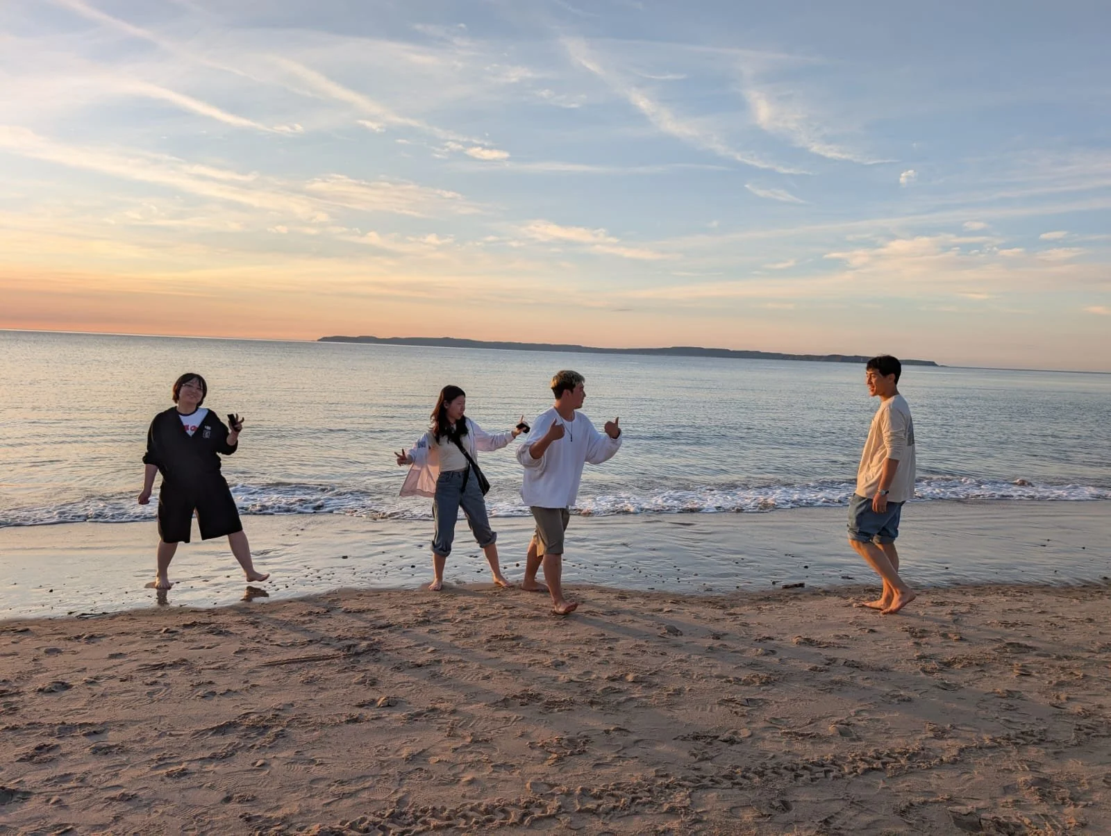 Five friends standing and walking along the beach at sunset, with the ocean and a distant landmass in the background.